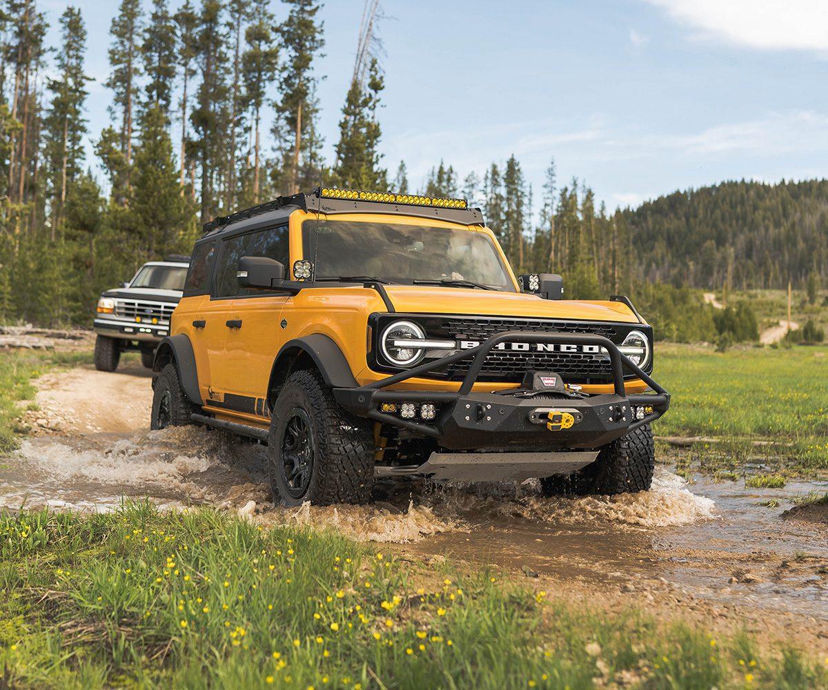 Yellow Ford Bronco with roof rack and off-road accessories driving through water on a forest trail