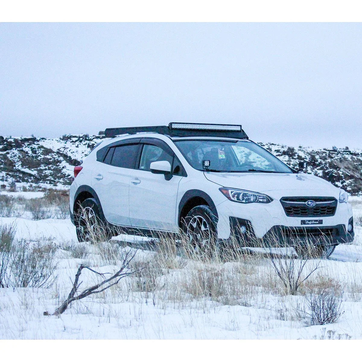 White Subaru crossover with roof rack and LED light bar parked on snowy off-road terrain