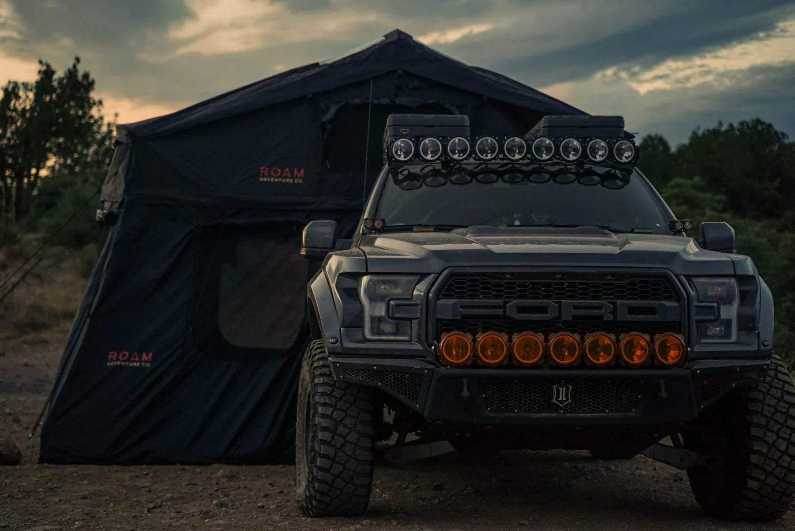 Ford off-road truck with roof lights parked by ROAM camping tent outdoors at sunset