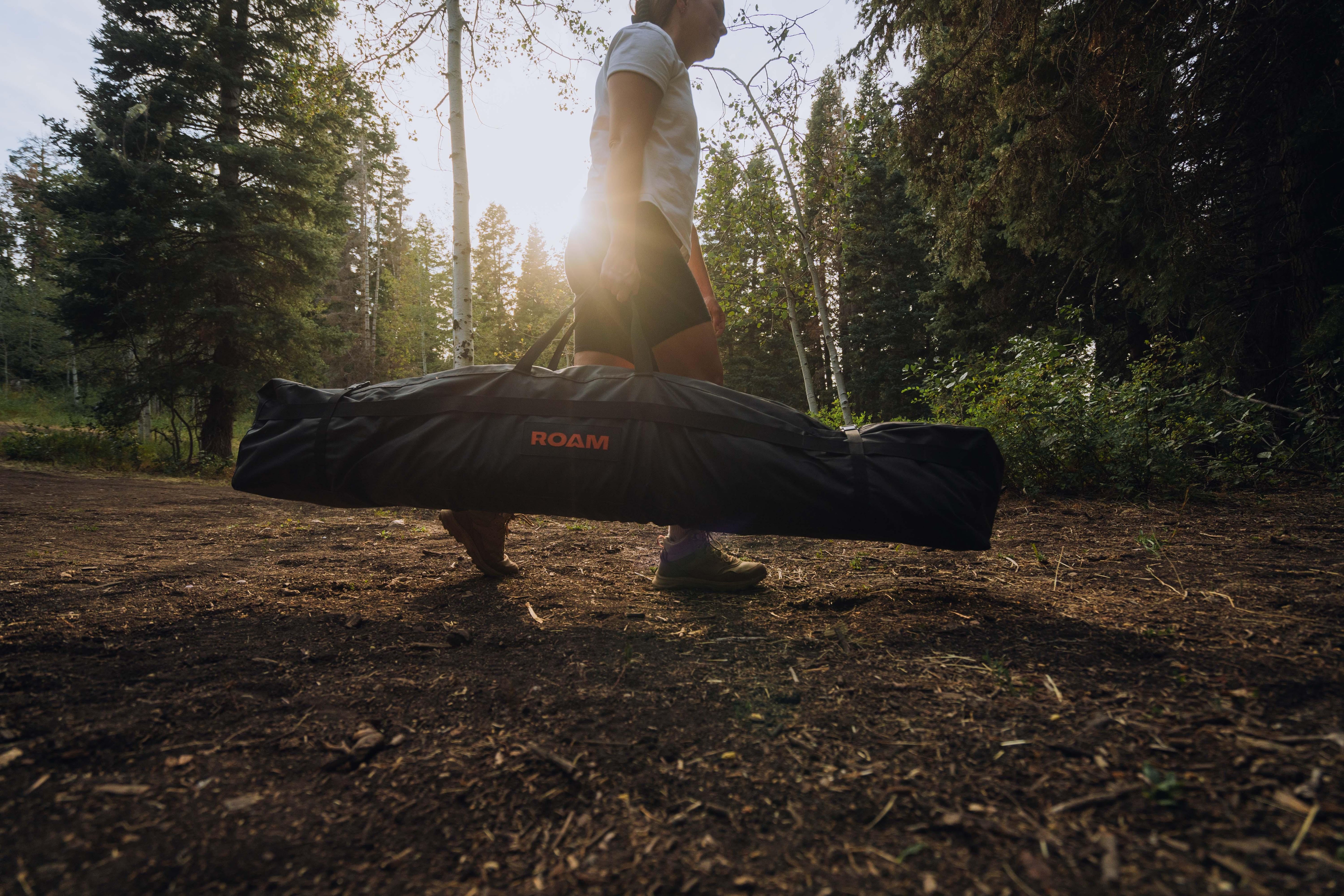 Person carrying a ROAM camping gear bag outdoors in a forest setting at sunset