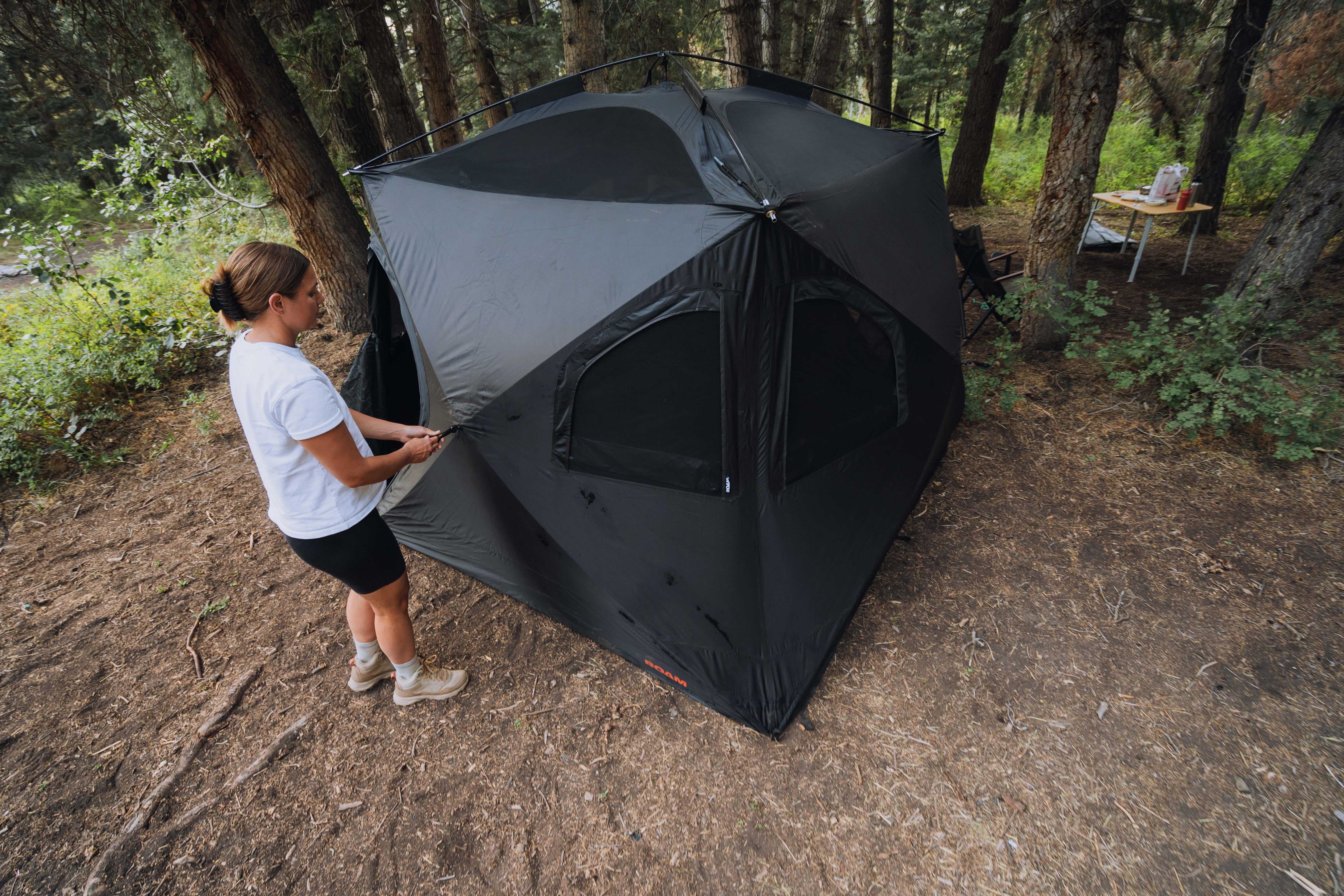 Woman zipping up a black, pop-up camping tent in a forest campsite with a picnic table nearby.