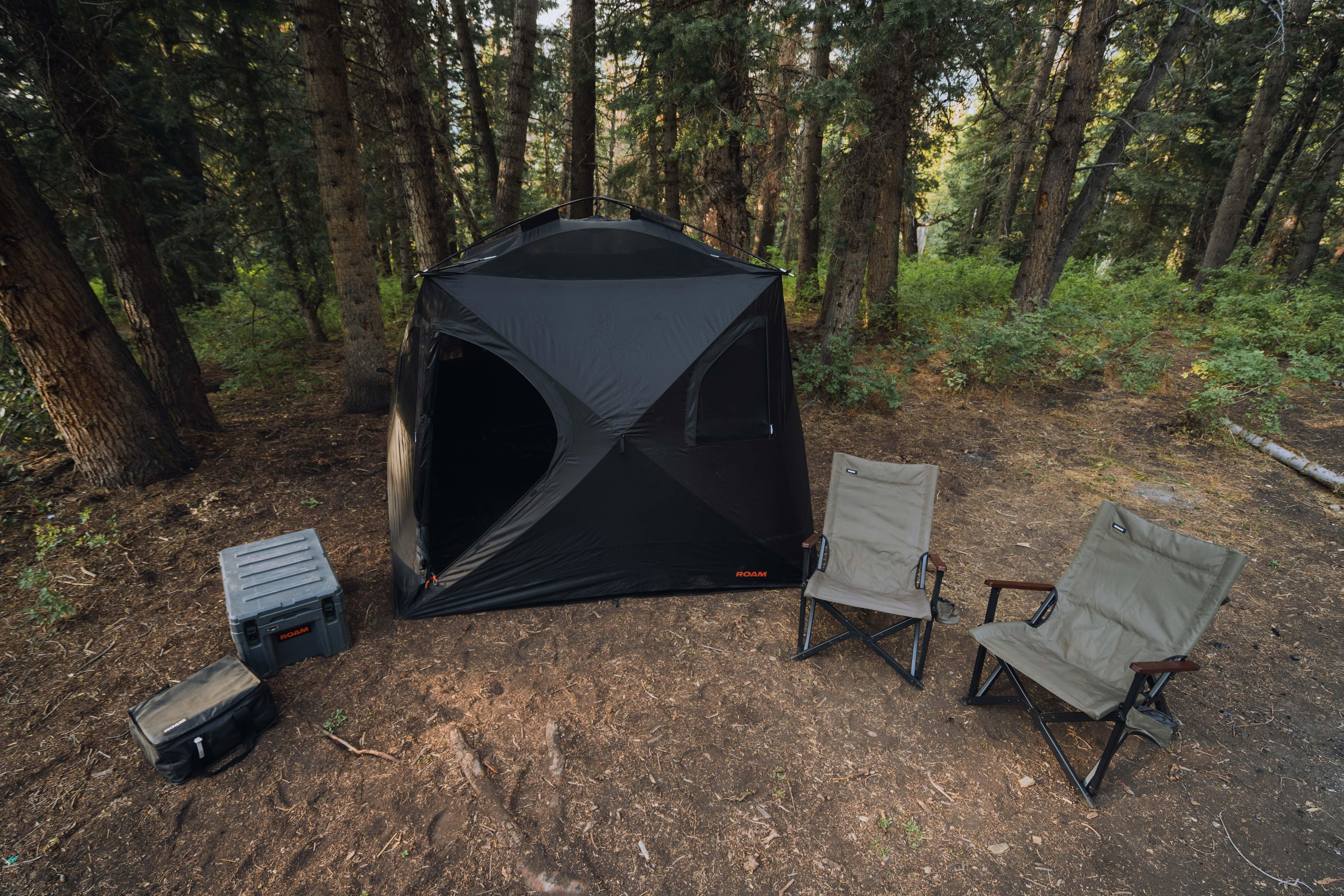 Overlanding campsite with black tent, camping chairs, and storage gear in forest setting