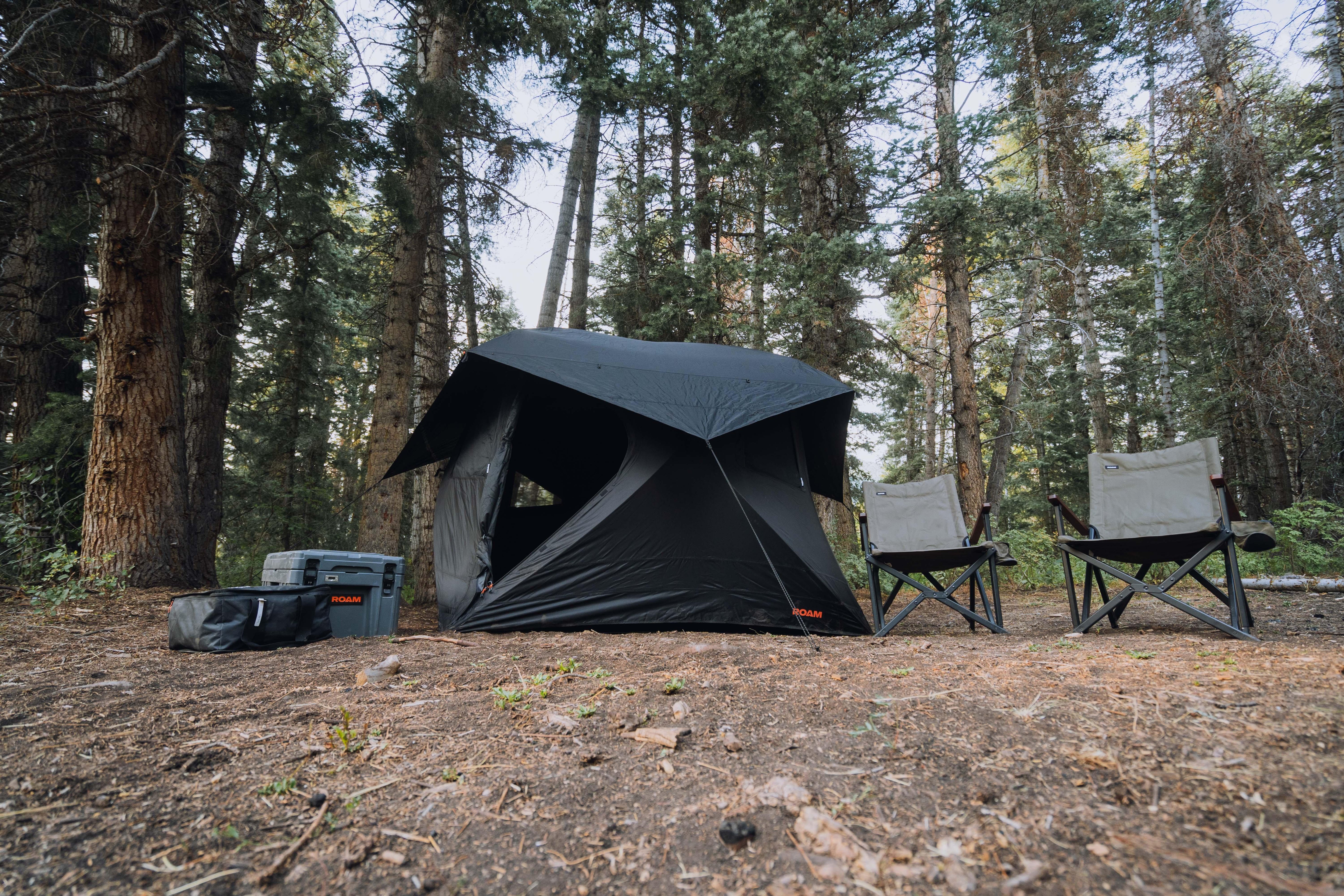 Overlanding camping setup with black tent, folding chairs, and gear in forest