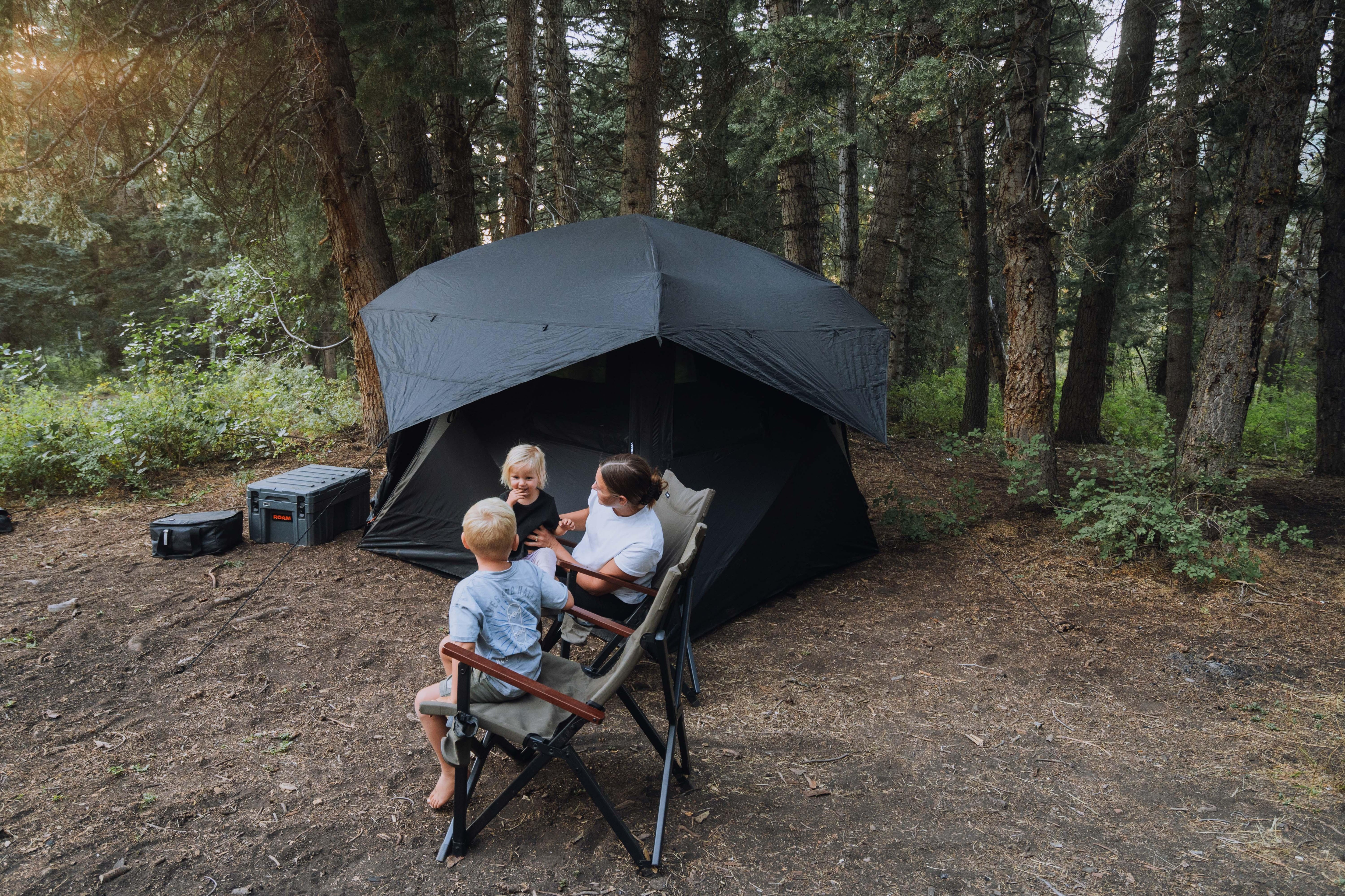 Overlanding campsite with black tent, gear boxes, and family relaxing in chairs among trees
