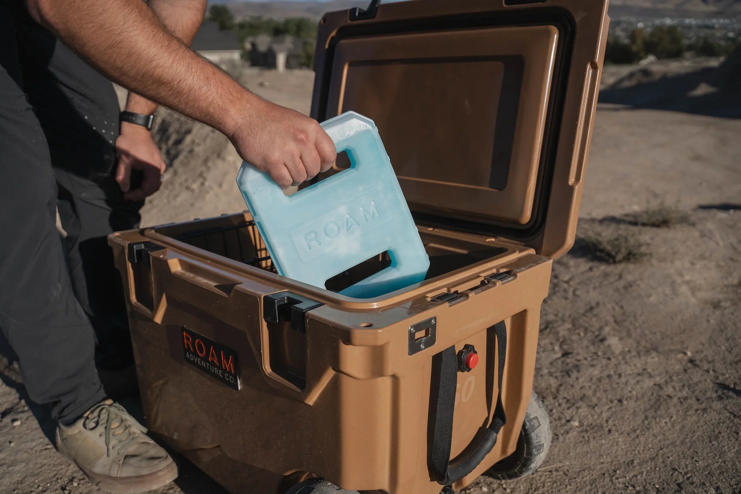 Person placing ice pack inside large tan outdoor cooler for camping or overlanding