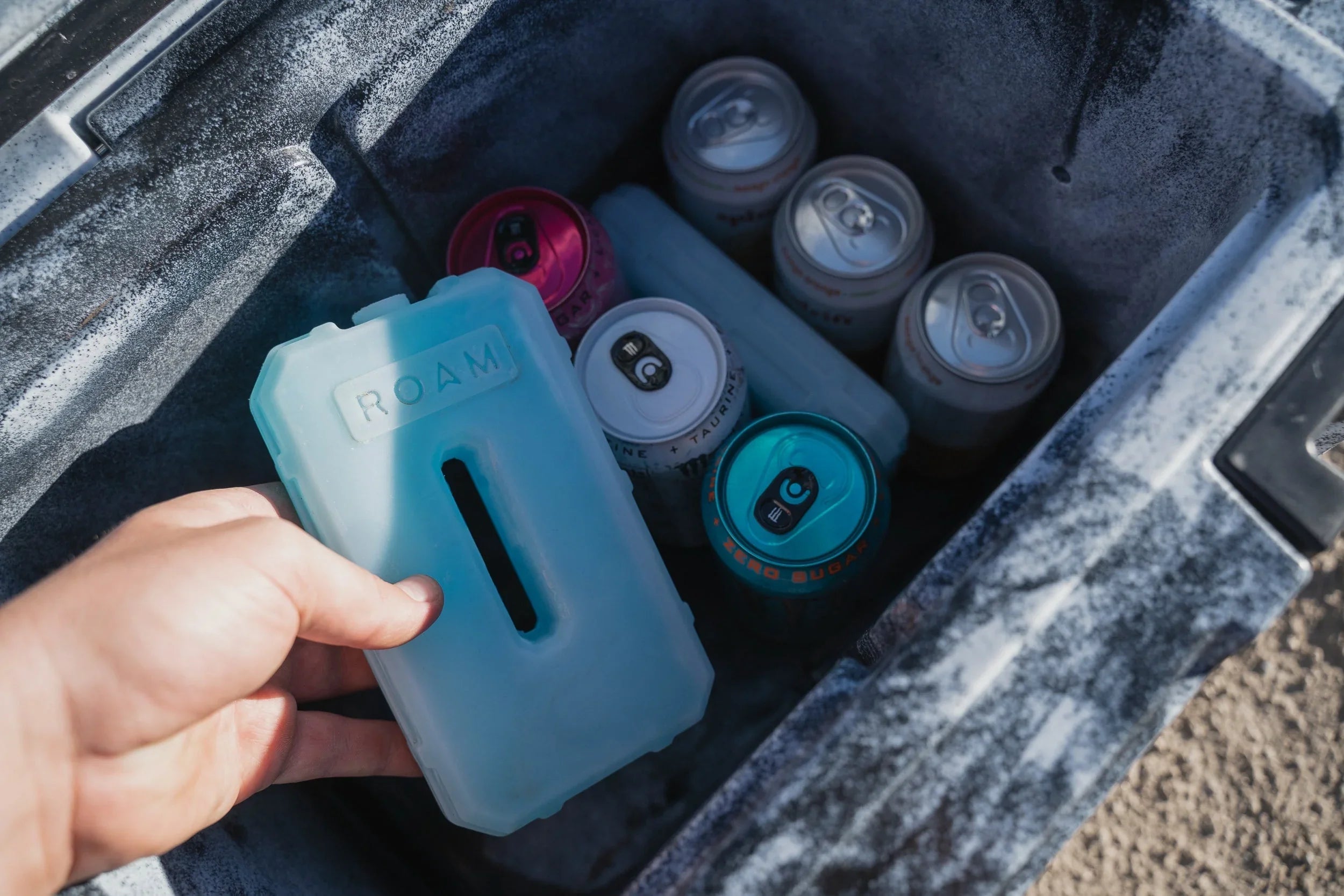 Hand placing blue ice pack in cooler with assorted canned drinks for overlanding adventure