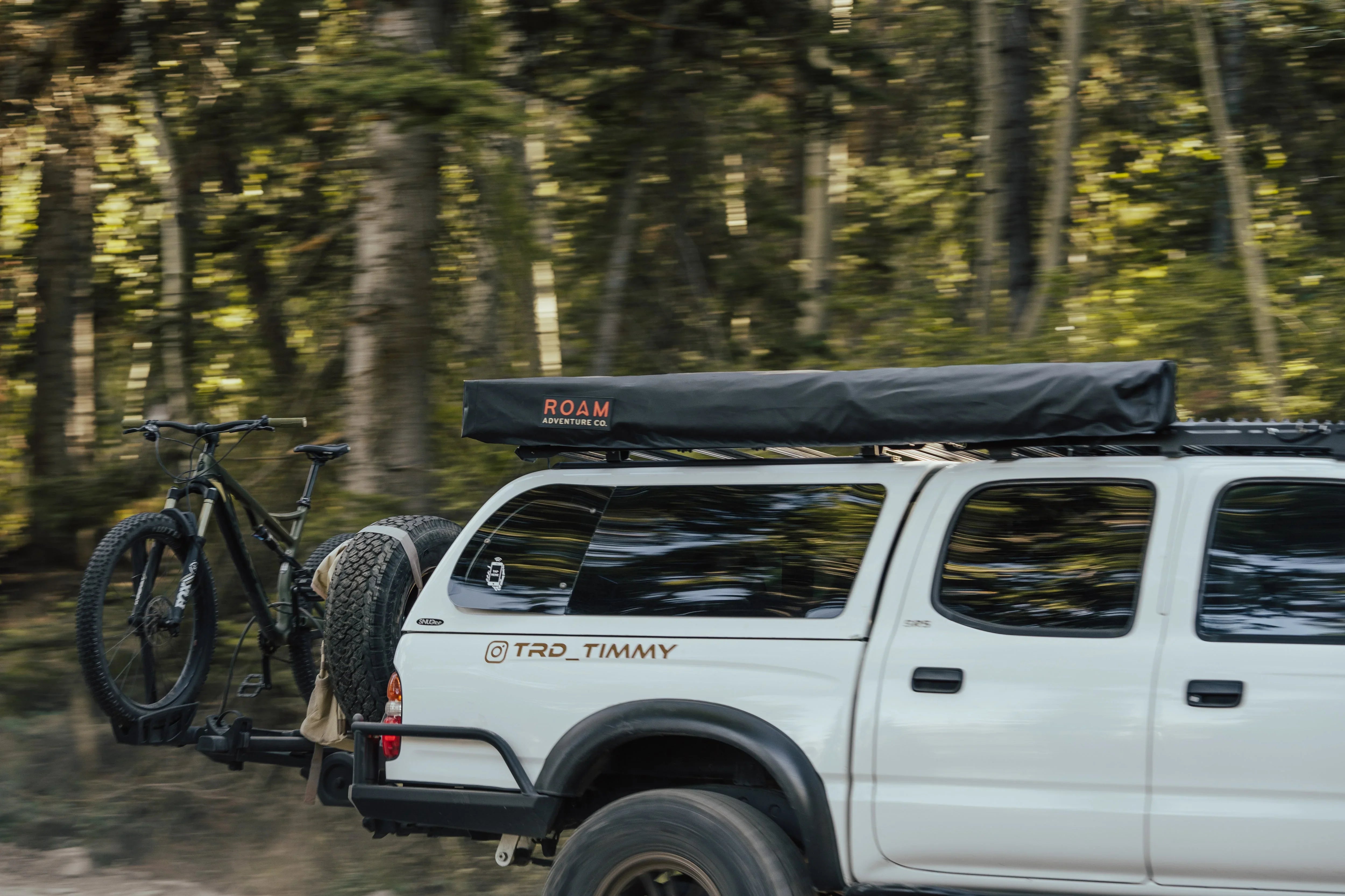 Toyota Tacoma with rooftop tent, spare tire, and mountain bike on forest trail