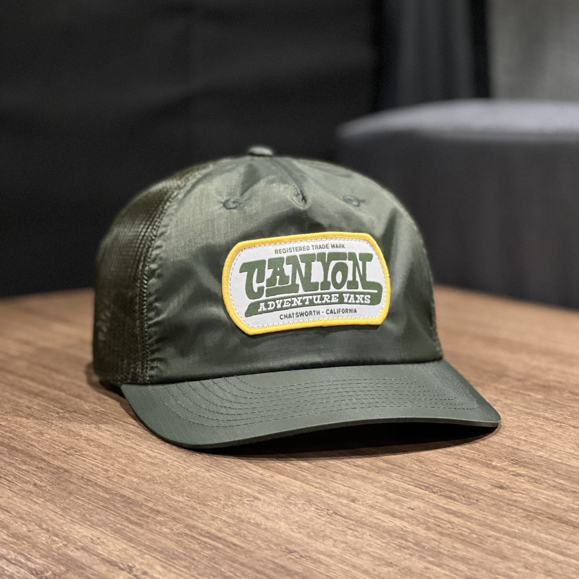 Olive green Canyon Adventure Vans trucker hat with mesh back and front logo patch on wood table