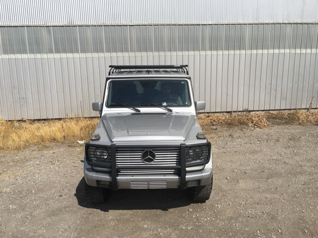 Silver Mercedes-Benz SUV with roof rack parked on gravel near metal building, overlanding ready