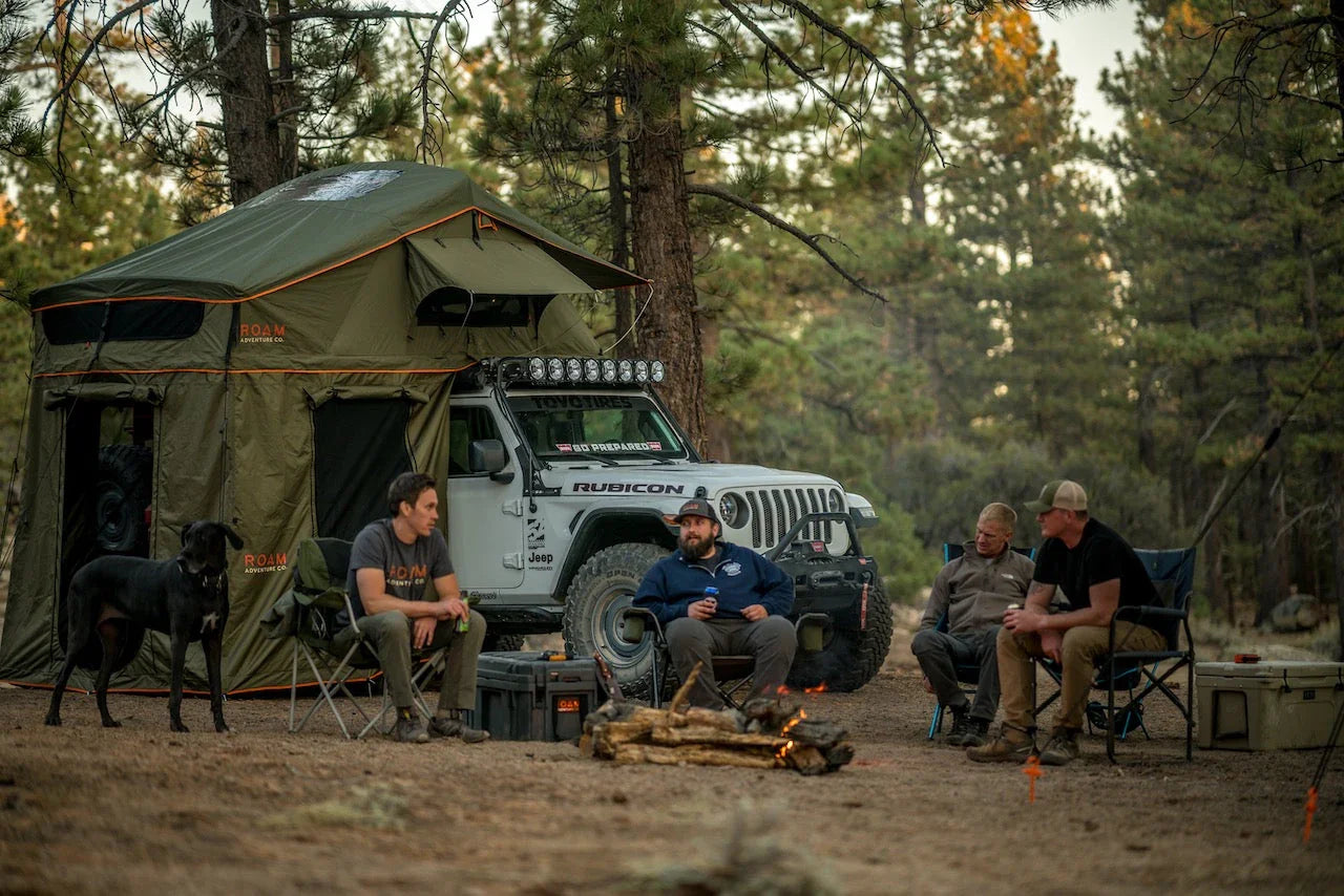 Group camping by Jeep Wrangler with rooftop tent in pine forest, overlanding gear visible