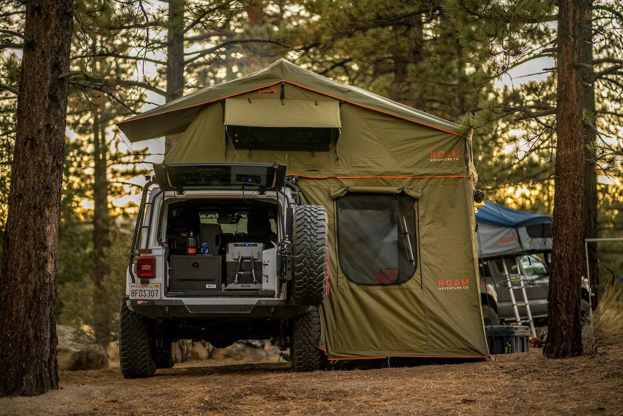 Overland vehicle with rooftop tent and camping gear set up in a pine forest at sunset