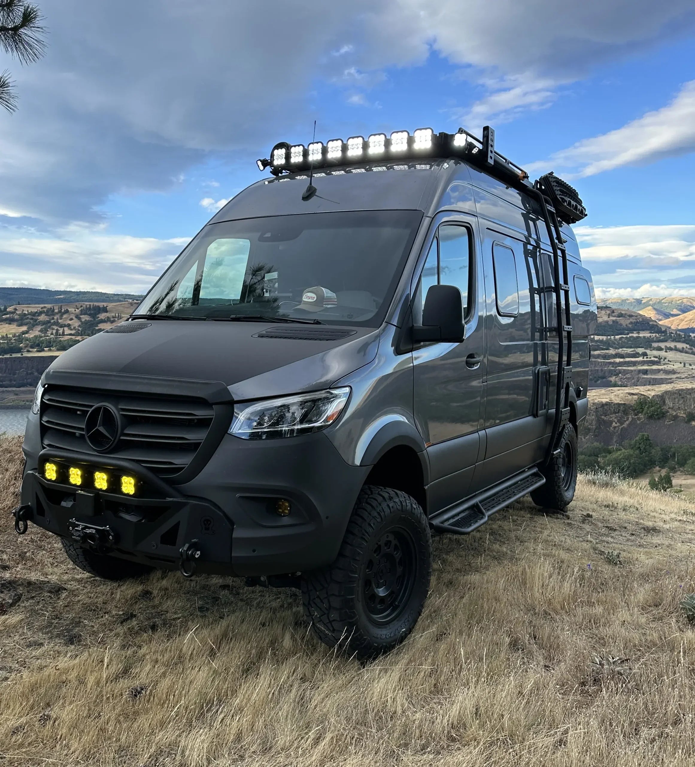 Off-road expedition van with roof rack, ladders, LED lights parked on grassy hillside