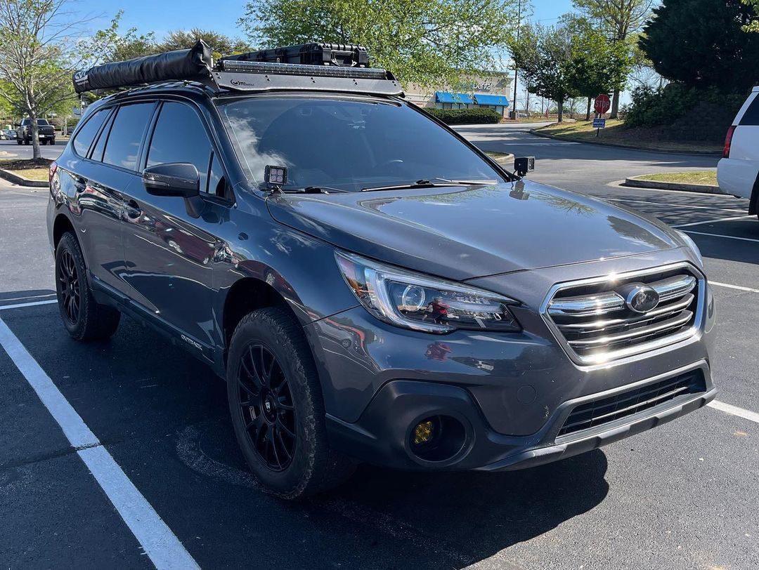 Gray Subaru Outback with off-road tires, roof rack, and mounted awning in parking lot