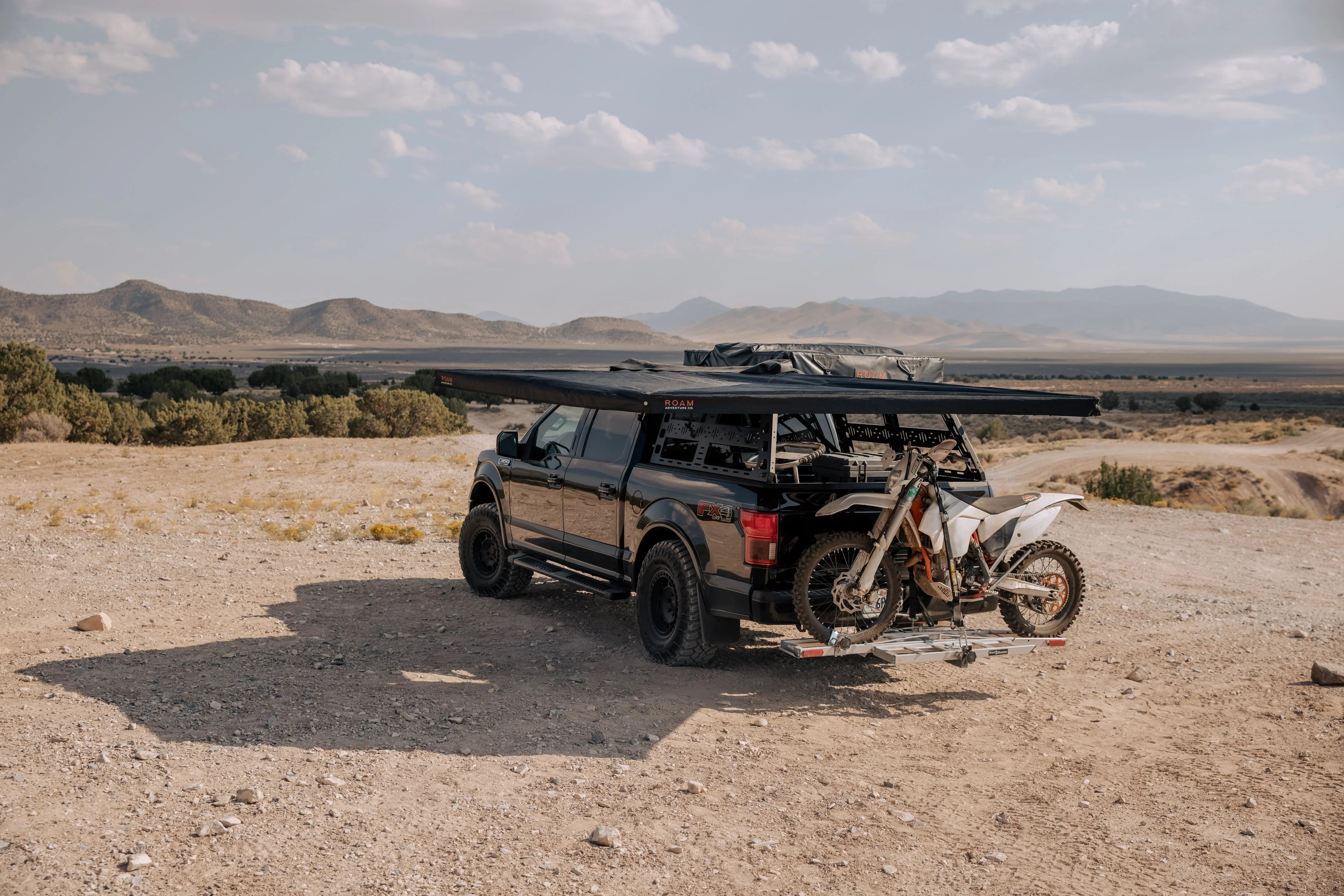 Overlanding truck with roof rack, awning, and dirt bike on hitch in desert landscape