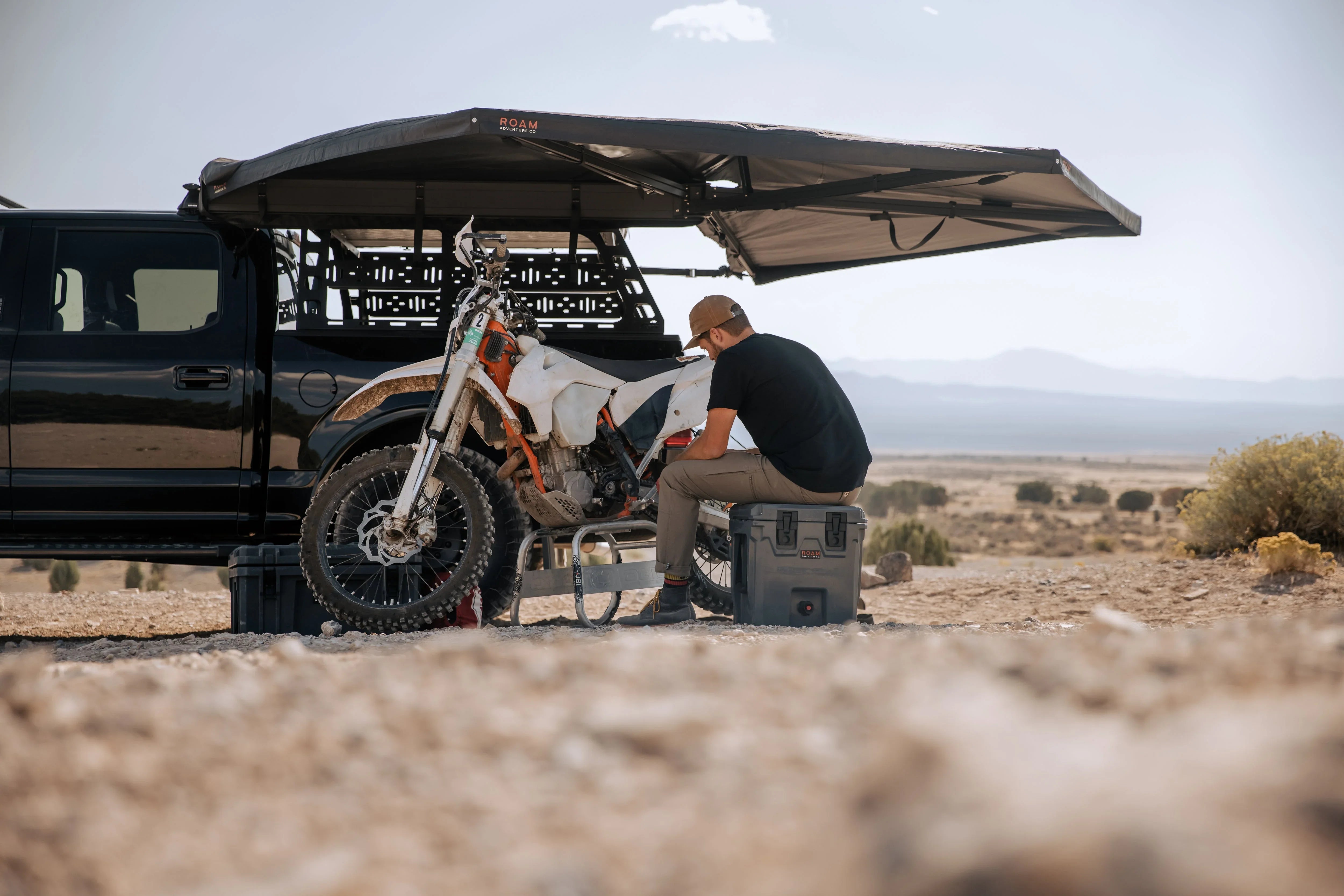 Man sitting under truck awning with dirt bike and rugged gear in desert camping scene