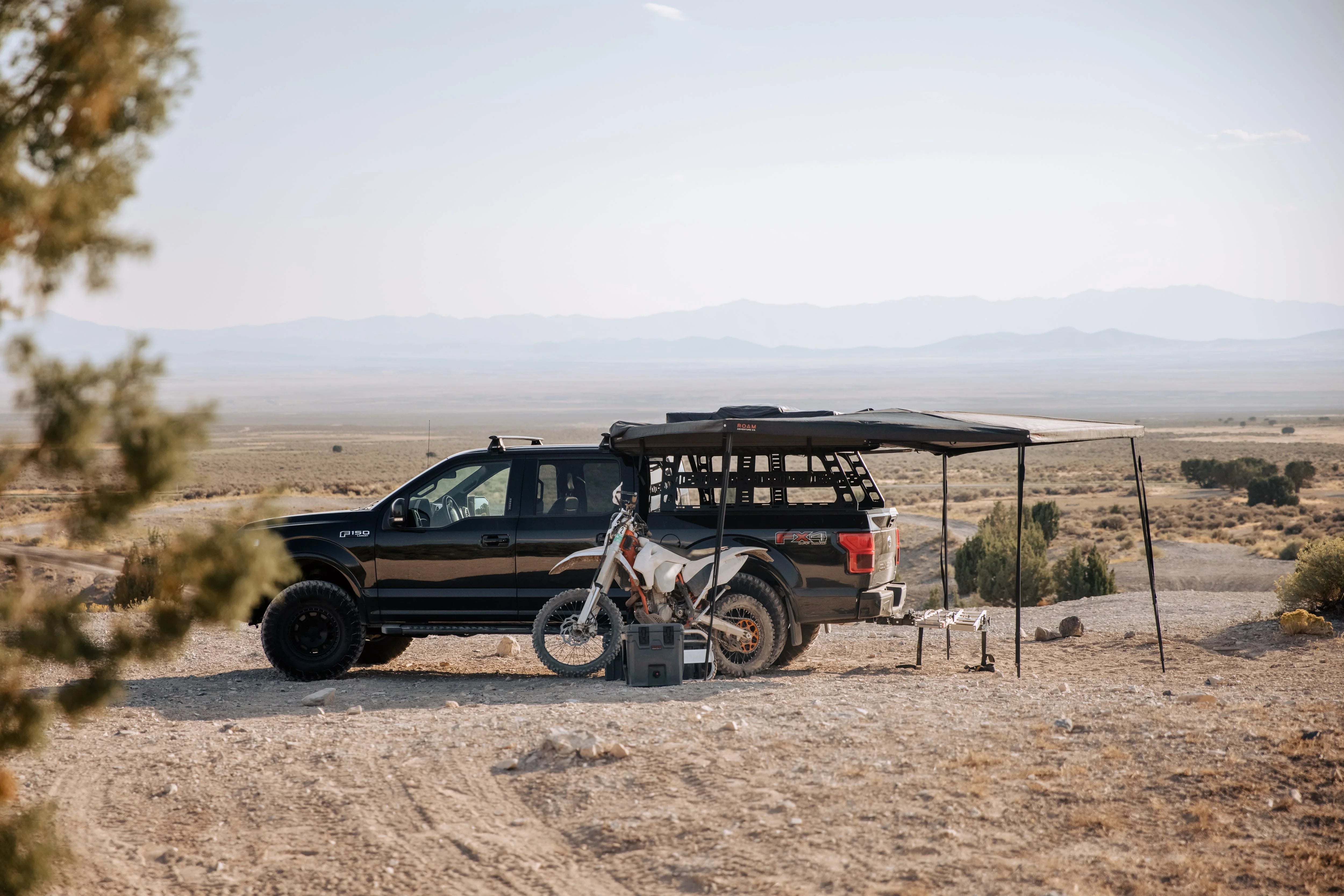 Overlanding truck with roof rack, awning, and dirt bike parked in a desert landscape