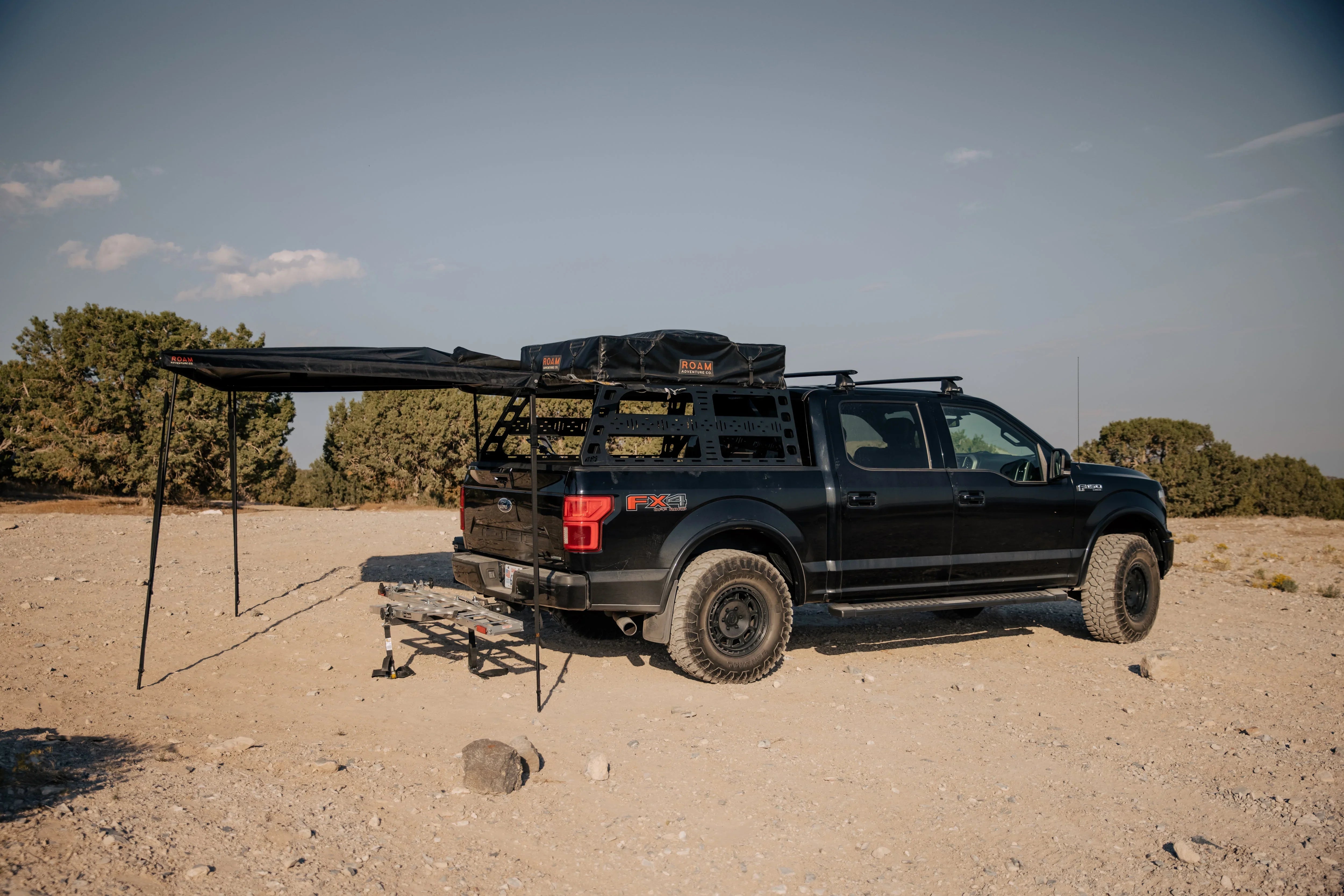 Black overland pickup truck with roof rack, rooftop tent, and awning in desert landscape