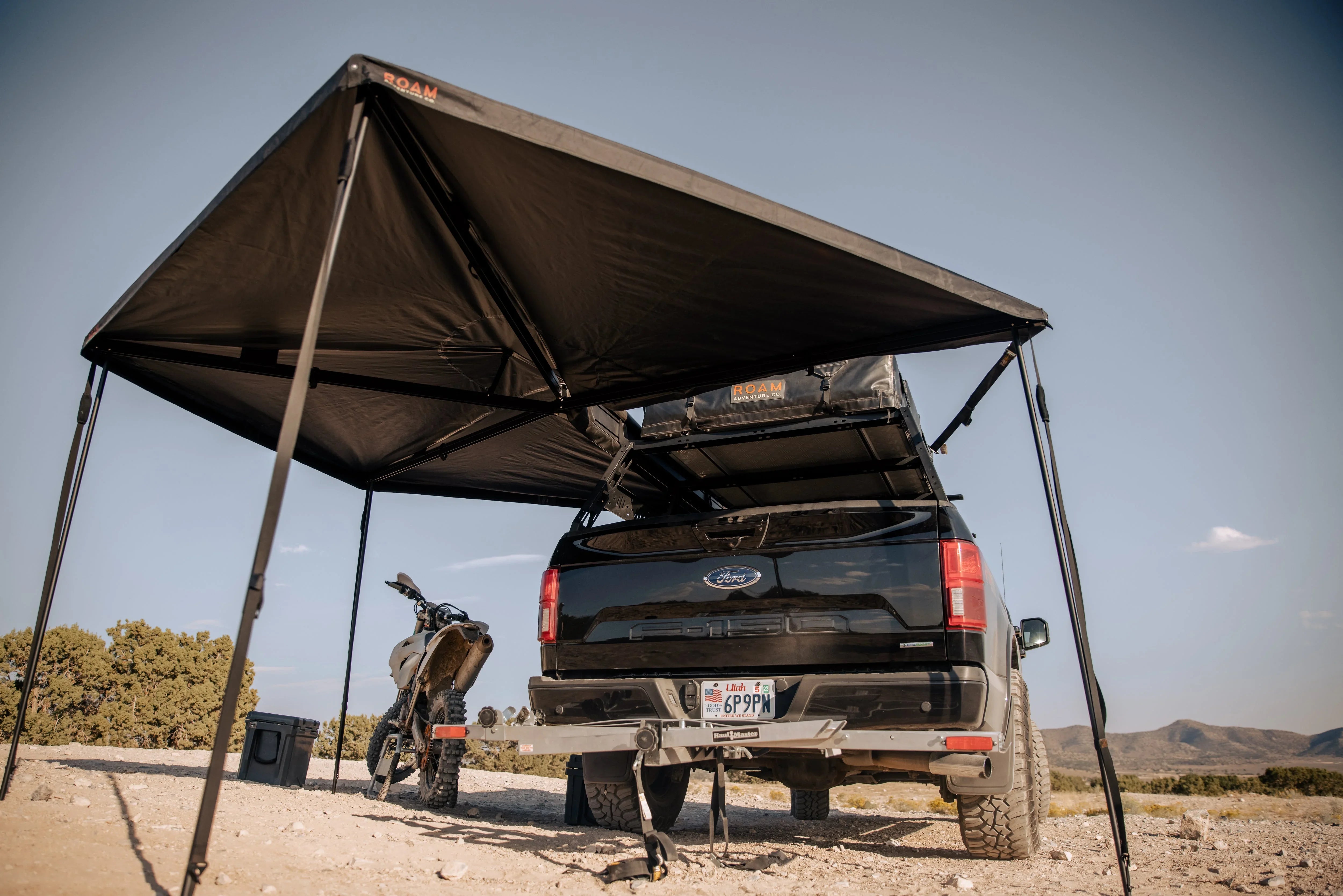 Overland truck with rooftop tent and awning setup, motorcycle parked beside in desert