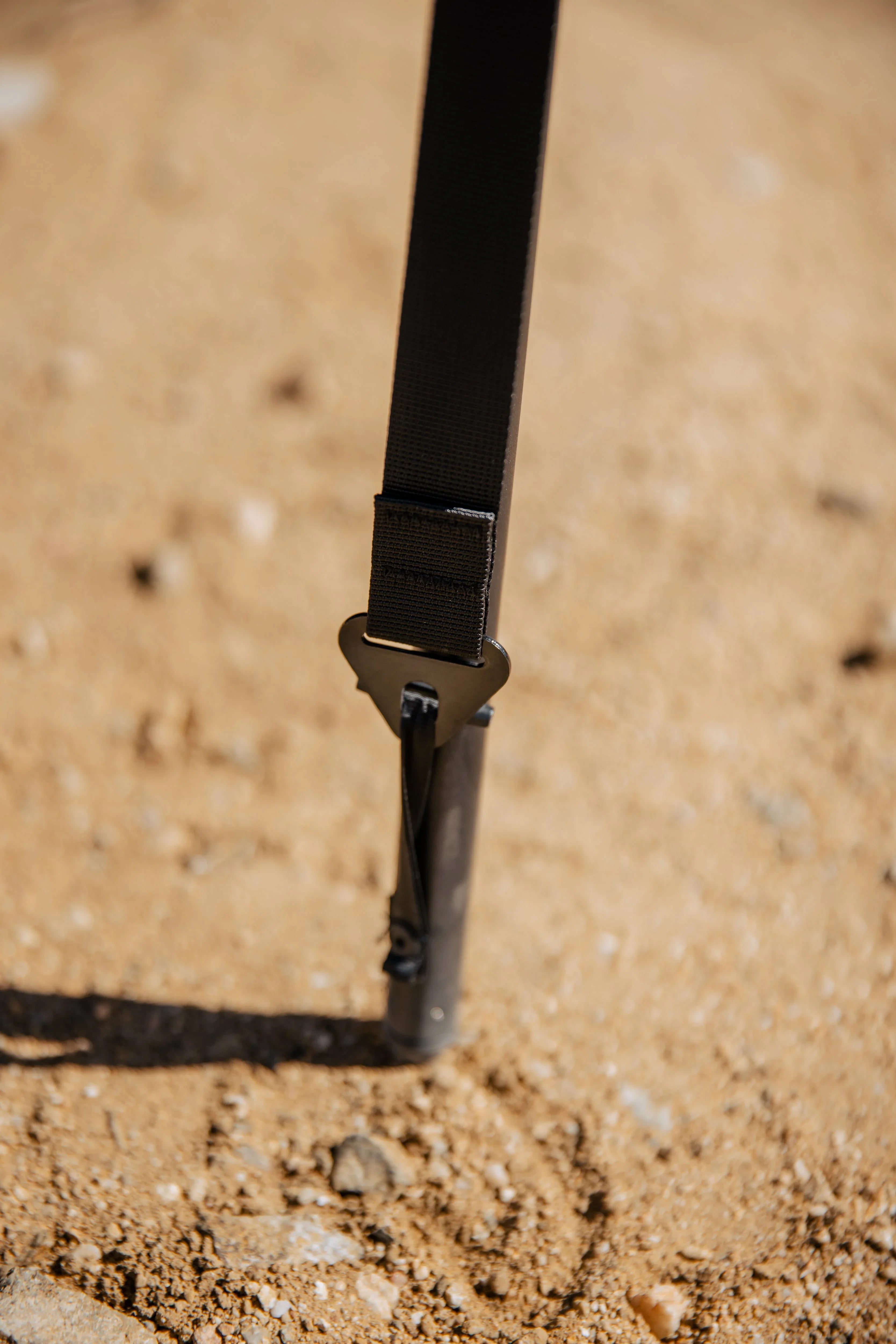 Close-up of a black camping strap and stake secured in sandy ground, outdoor gear detail.