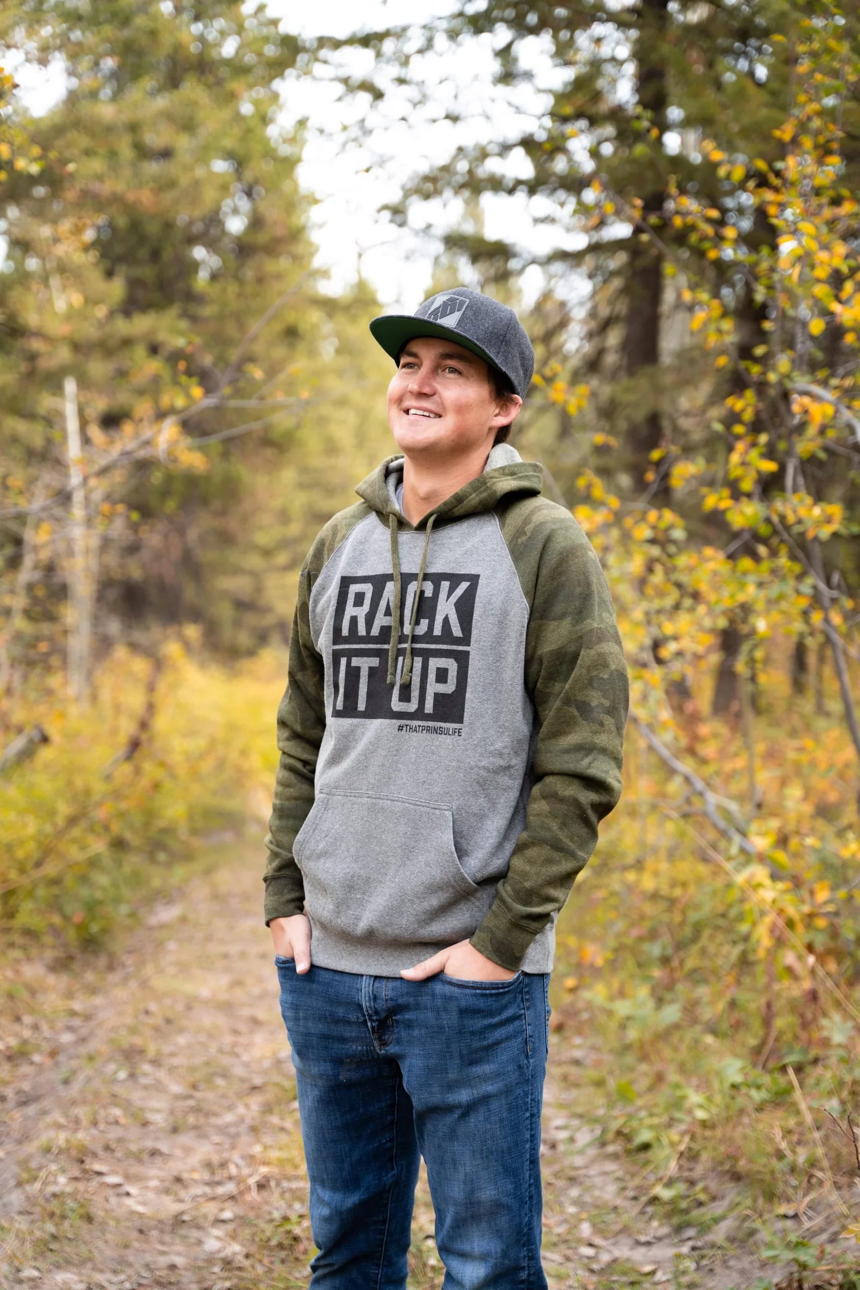 Man in 'Rack It Up' hoodie and cap standing on forest trail, outdoor adventure apparel