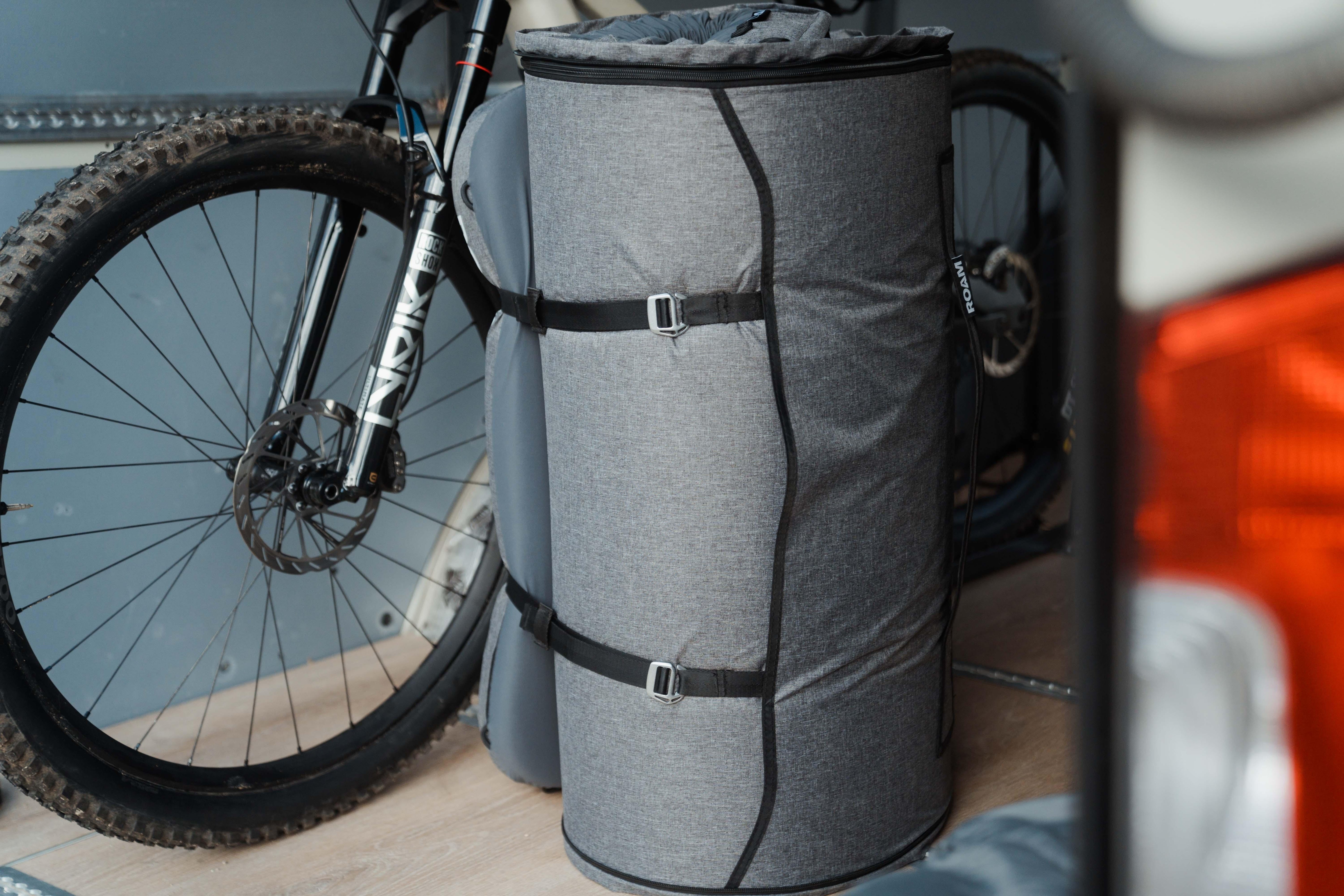 Gray roll-up camping mattress with straps, stored beside mountain bike indoors