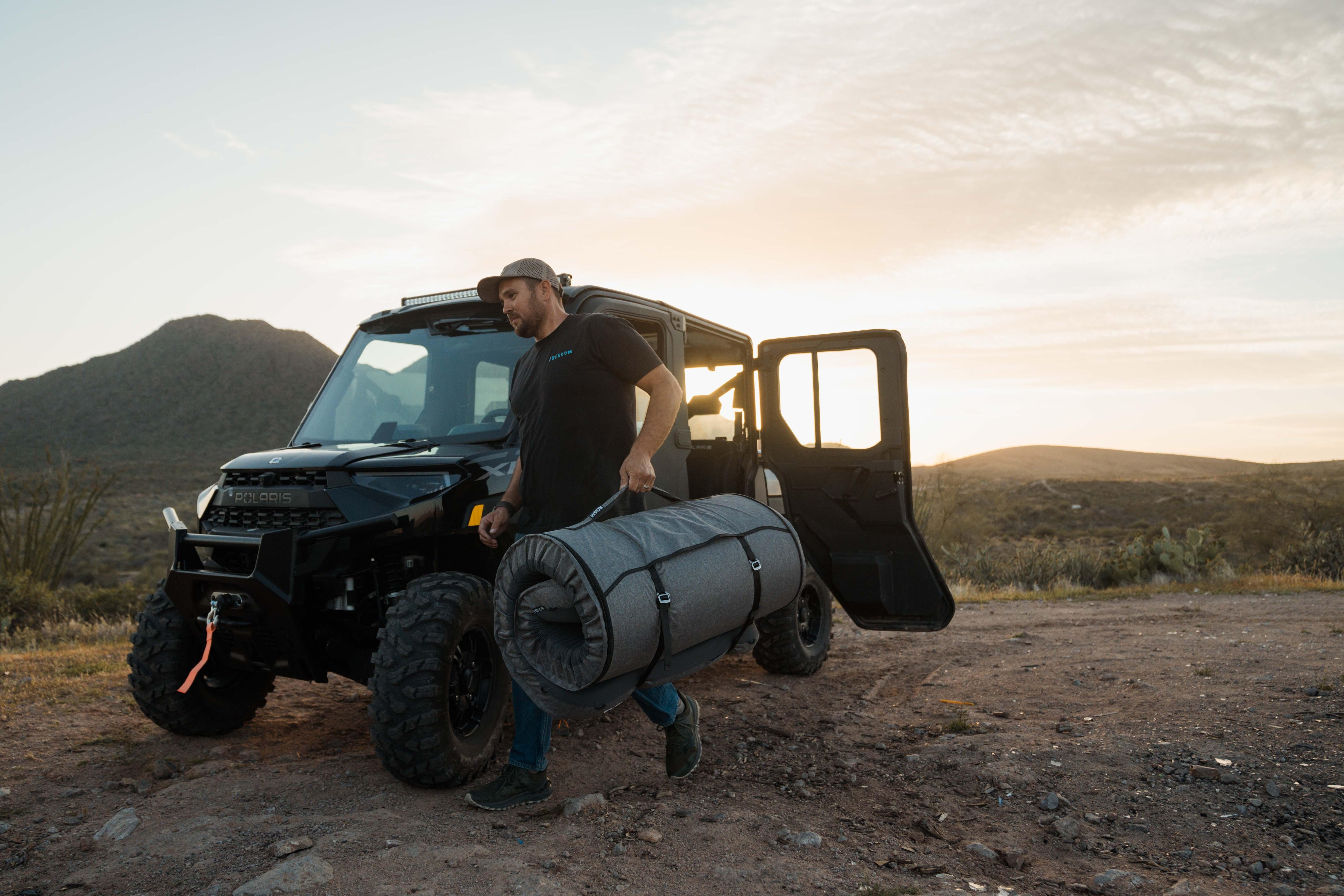 Man carrying rolled camping mattress near off-road vehicle at desert campsite at sunset