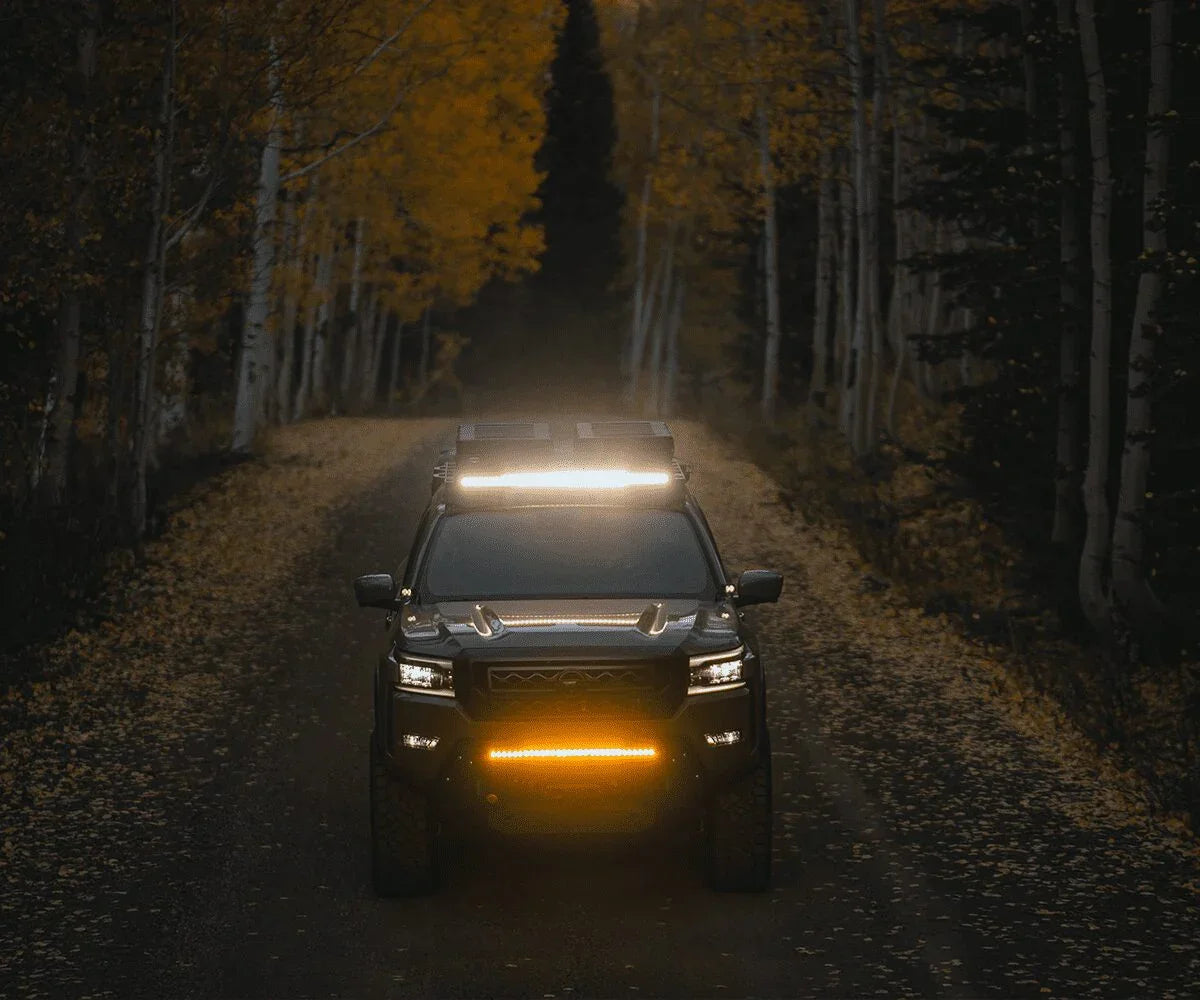 Nissan Frontier with roof rack and LED light bar driving on a forest trail at dusk