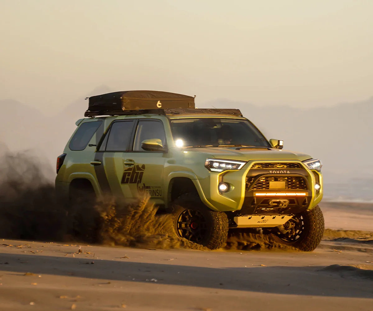 Toyota 4Runner with off-road bumper driving on sandy beach at sunset, overlanding gear visible
