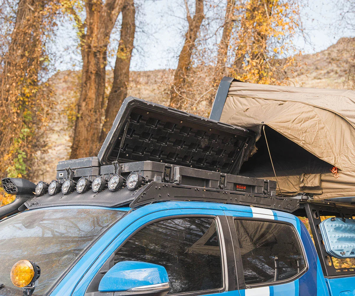 Blue Toyota Tacoma with overlanding roof rack, rooftop tent, storage boxes, and off-road lights