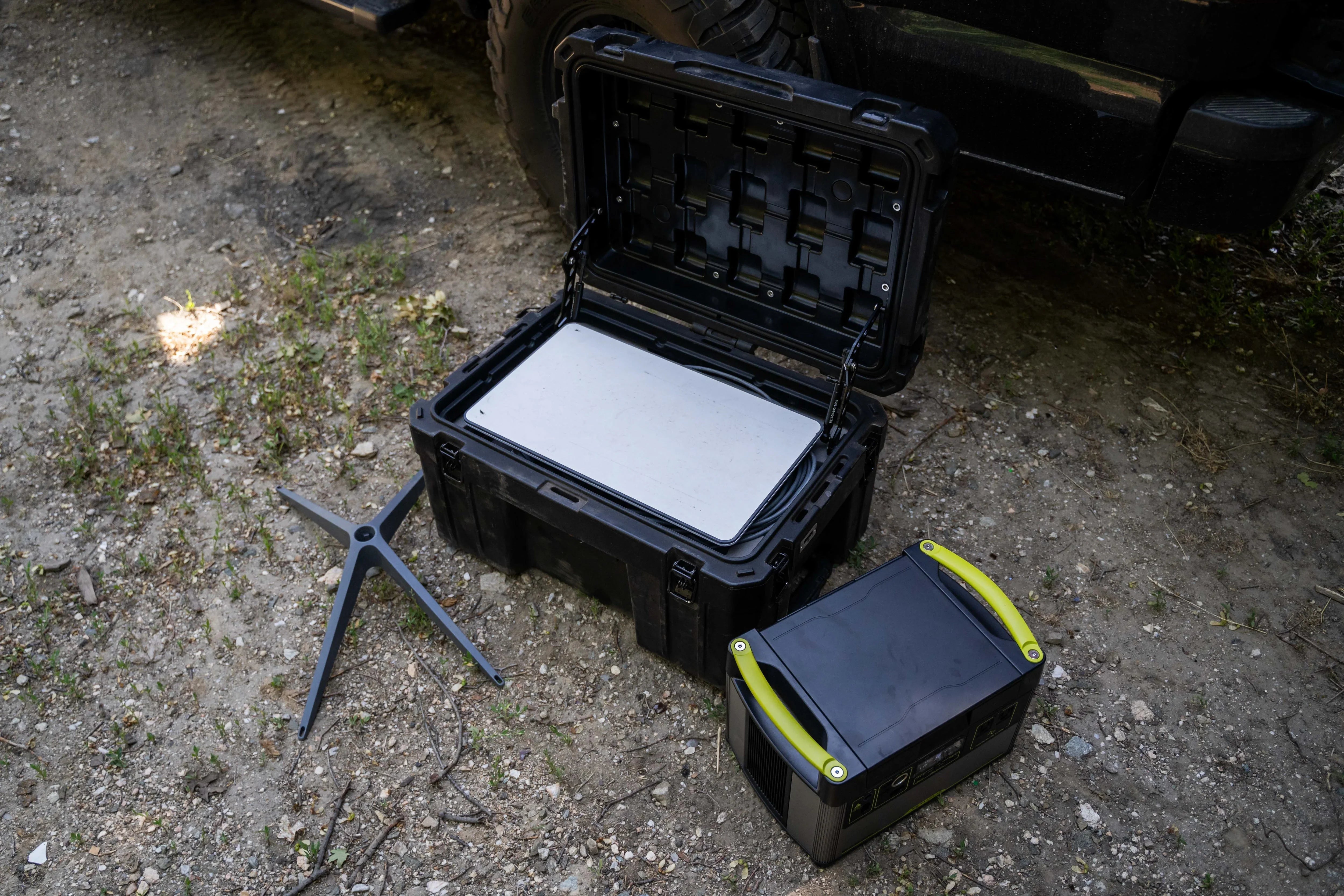Off-road portable power station, rugged storage box, and tripod stand on dirt next to a vehicle