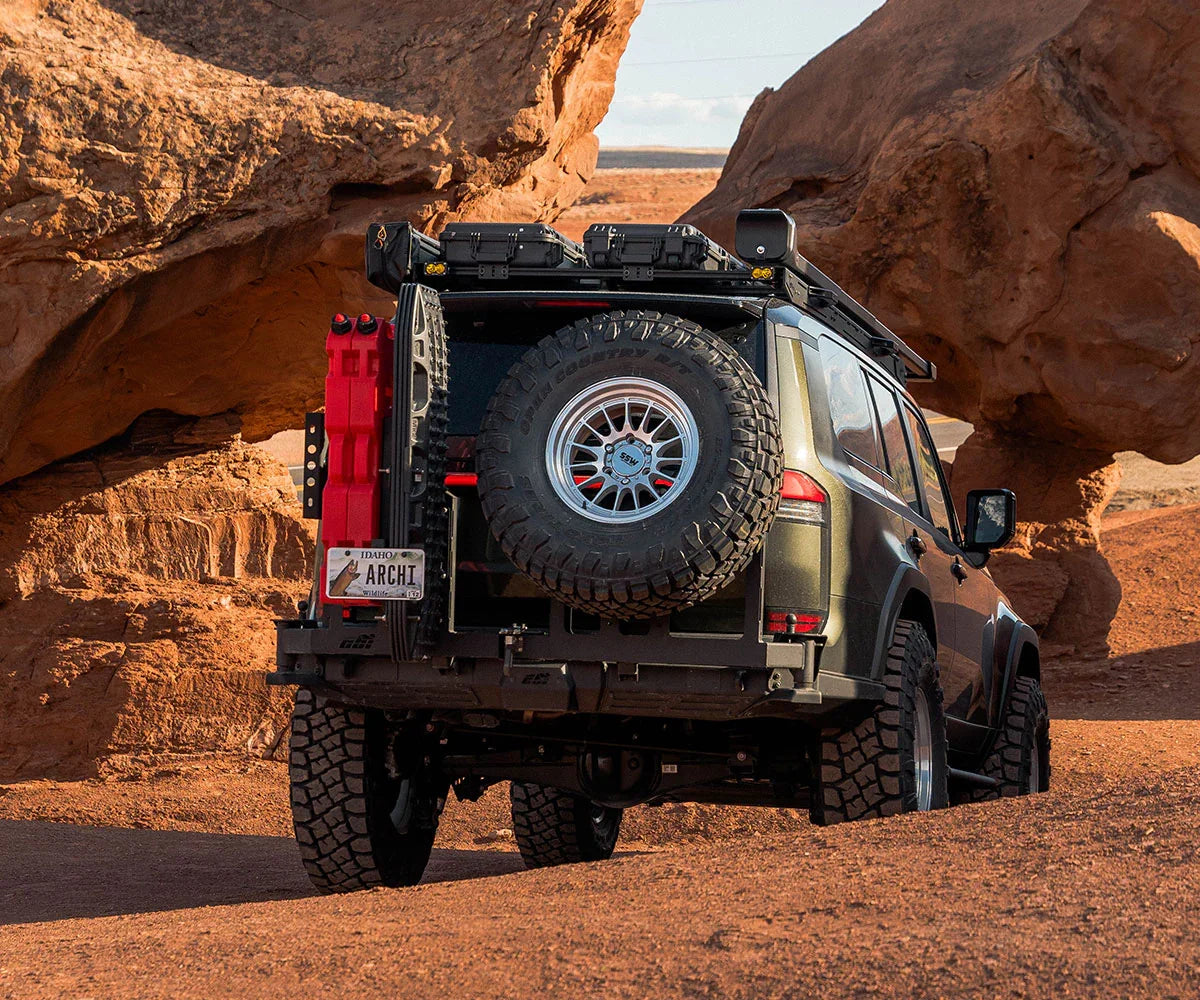 Off-road SUV with roof rack, large spare tire and gear, parked among desert red rocks