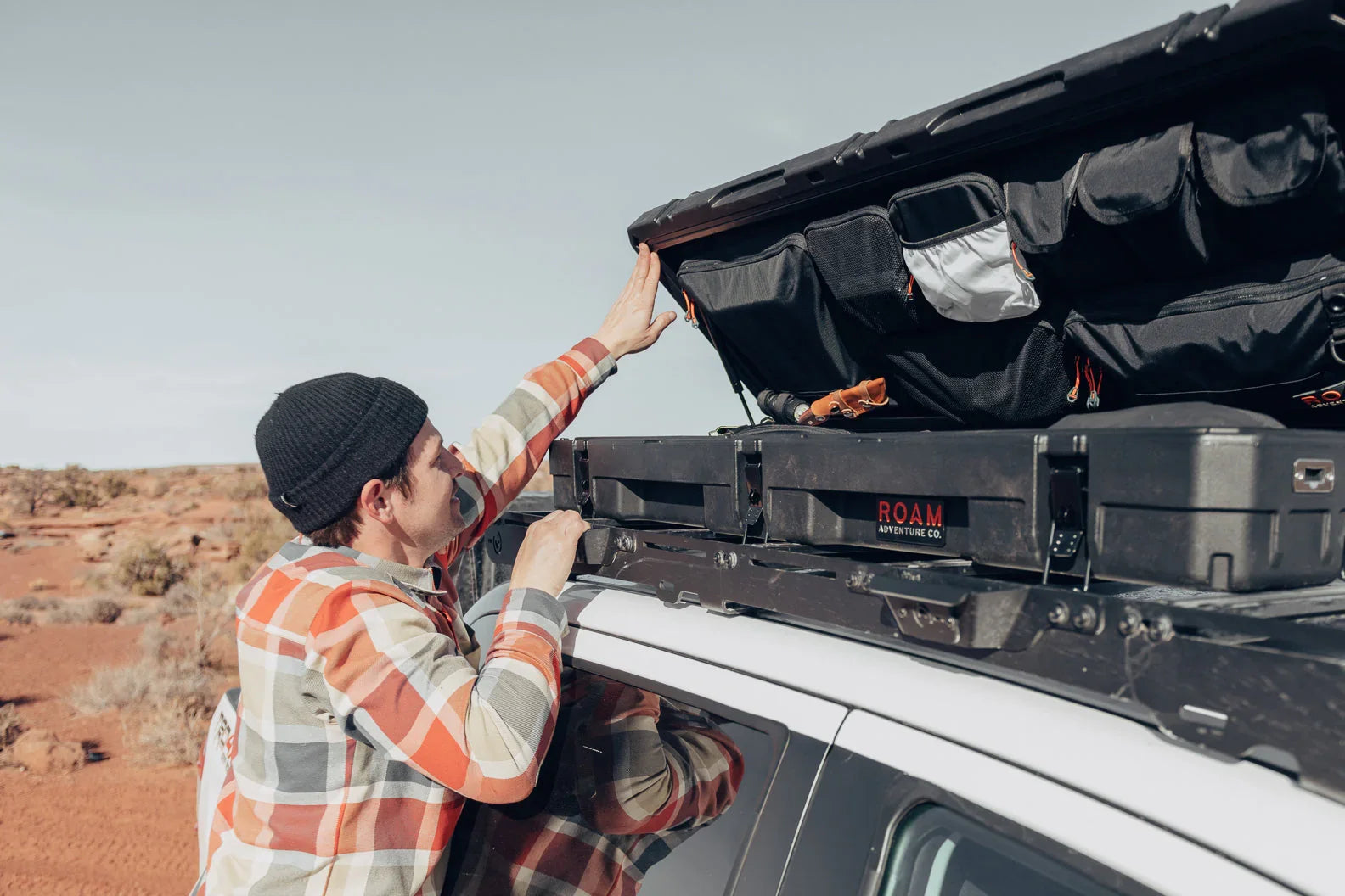 Man opening ROAM Adventure storage case on vehicle roof rack in desert landscape, overlanding gear