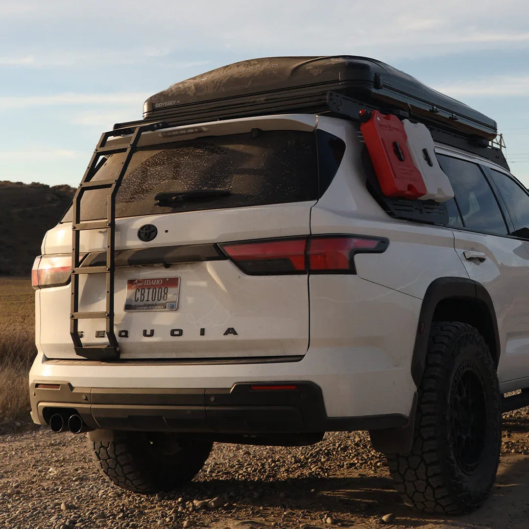 Toyota Sequoia SUV with roof rack, rear ladder, and off-road camping gear on gravel road