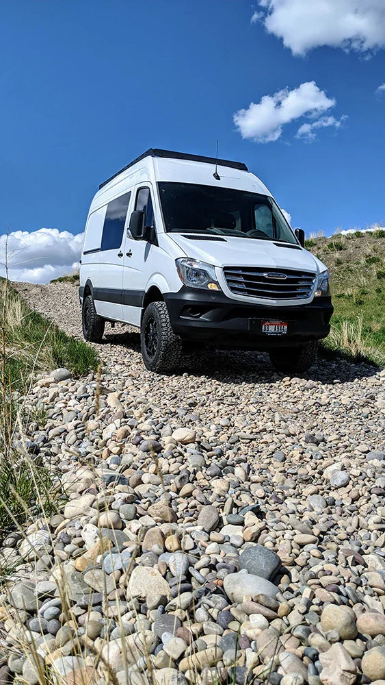White Sprinter van with roof rack on rocky off-road trail under blue sky