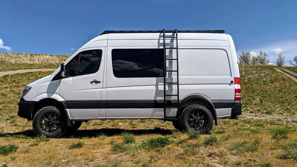 Sprinter van with roof rack and ladder parked on grassy terrain under blue sky