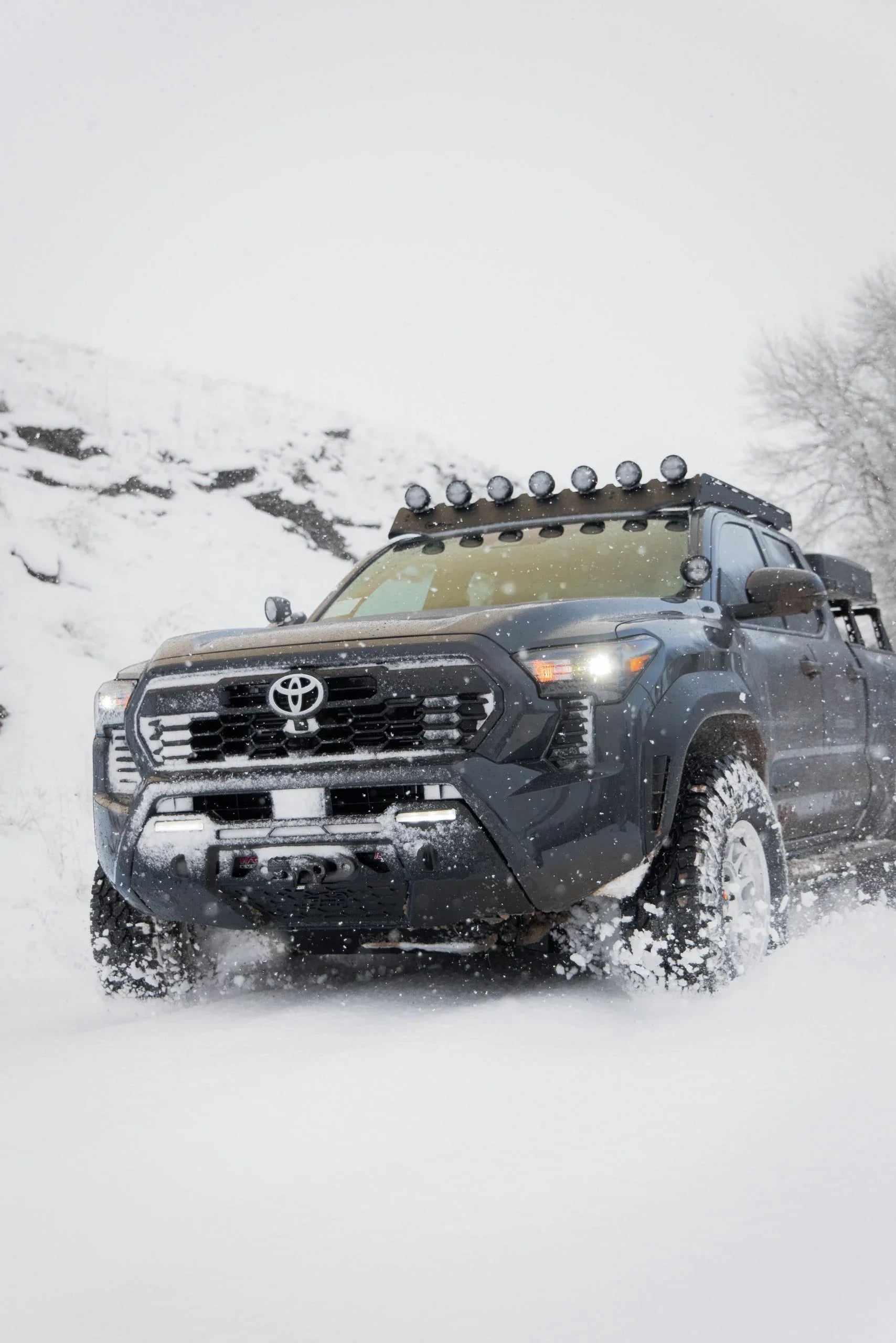 Toyota Tacoma with roof rack and off-road lights driving in snowy terrain