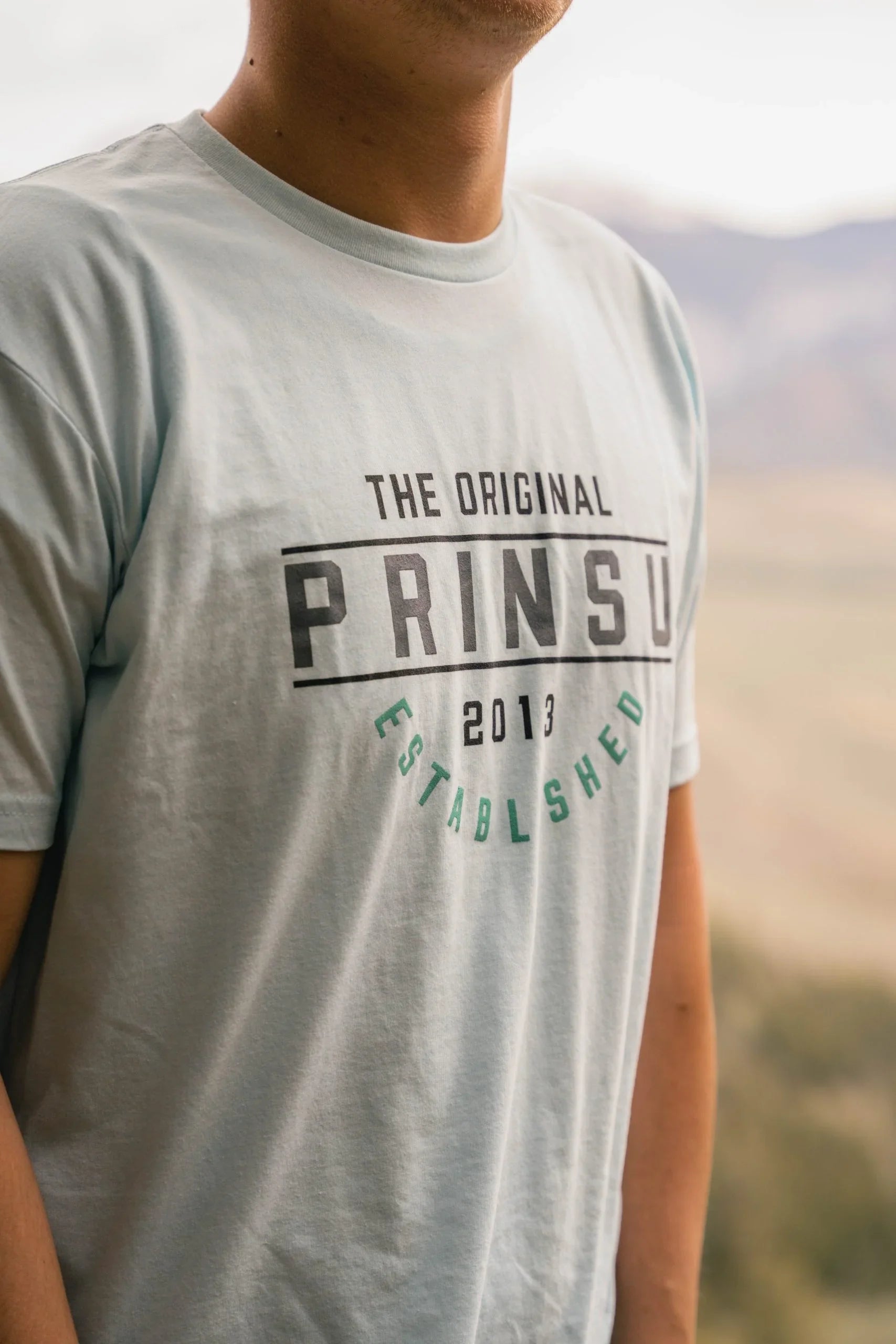 Person wearing light blue PRINSU t-shirt for overlanding and adventure in outdoor setting