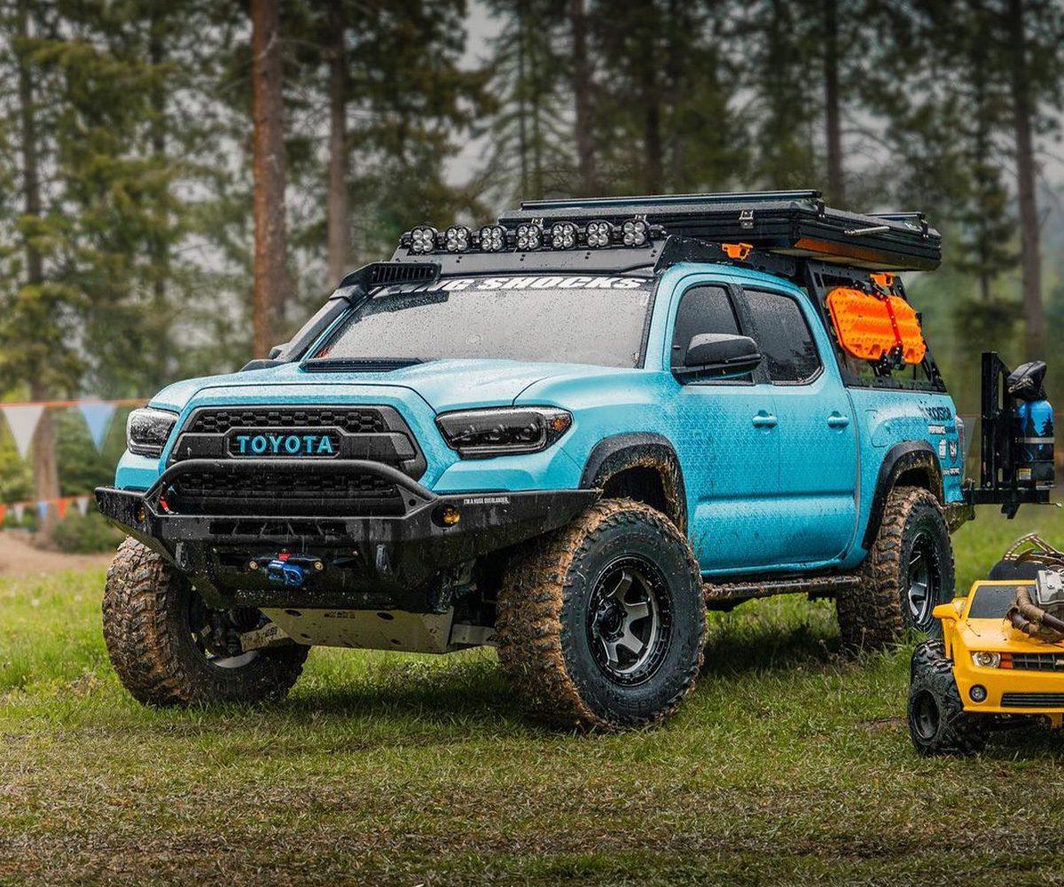 Lifted blue Toyota Tacoma with Prinsu roof rack and off-road gear in a forest setting