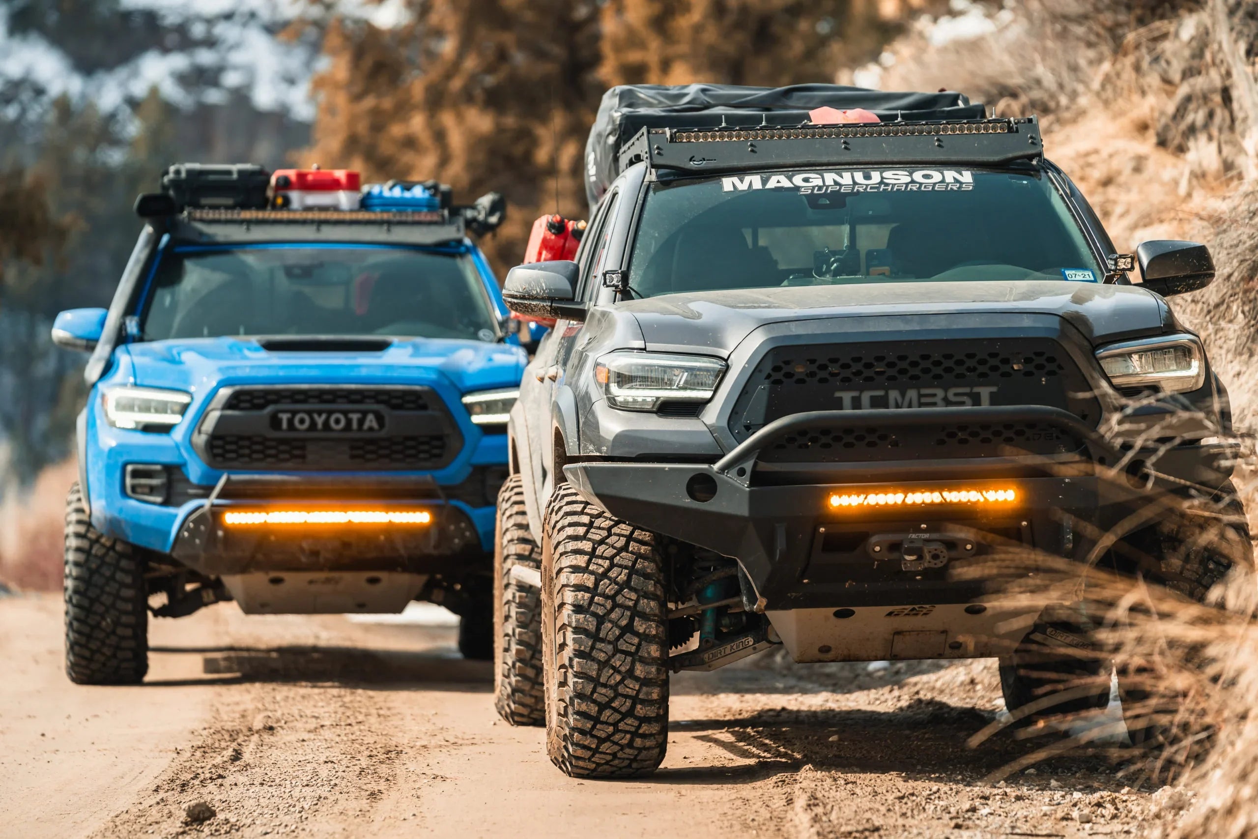 Two lifted Toyota Tacoma trucks with front bumpers and off-road accessories on a dirt trail