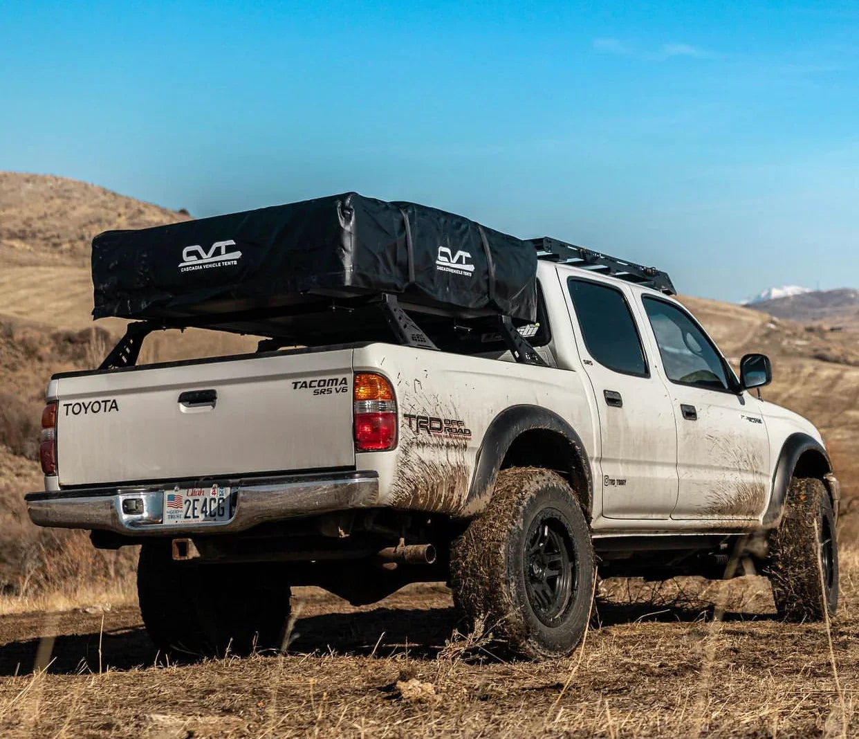 White Toyota Tacoma with overland bed bars and rooftop tent parked off-road in a hilly landscape