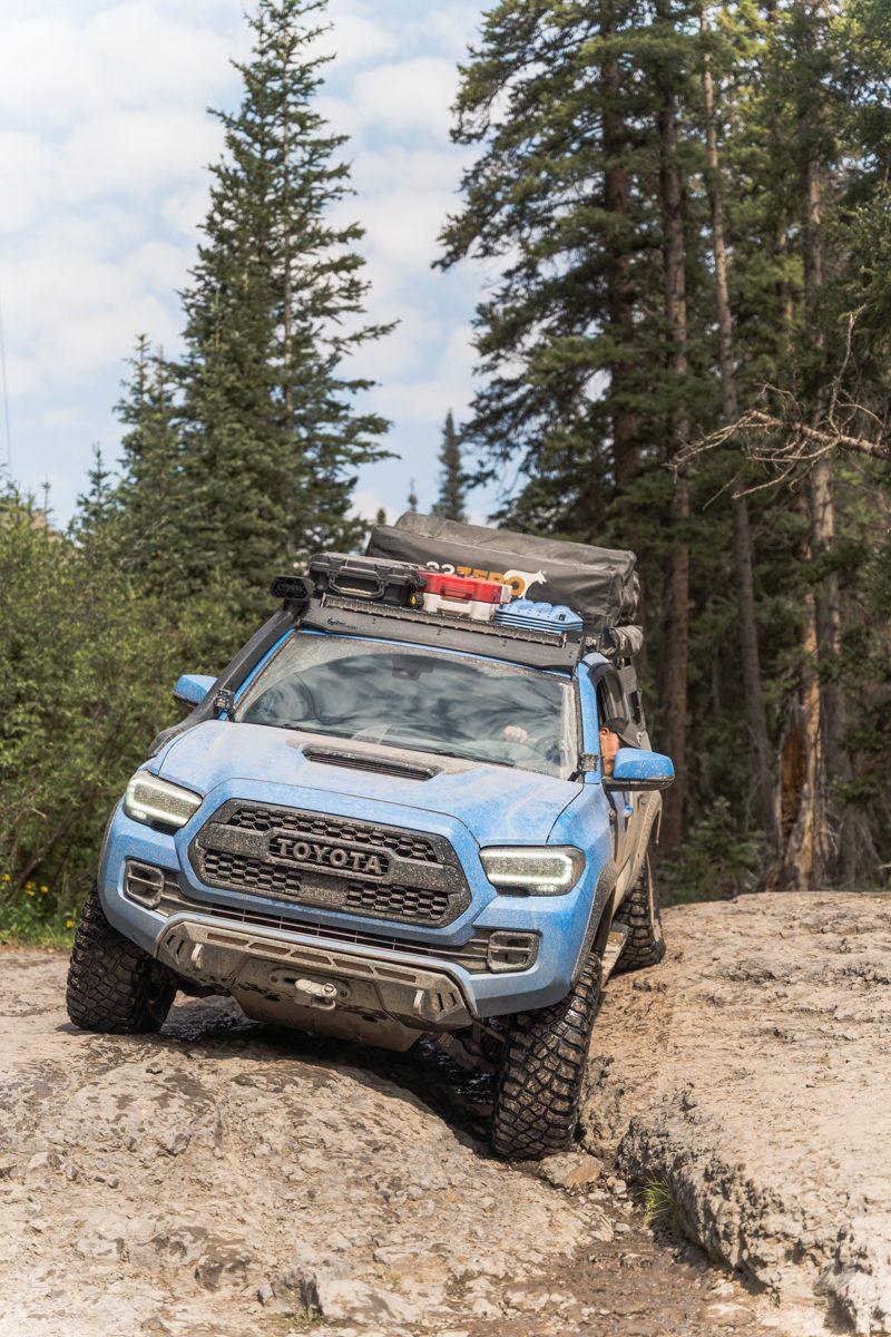 Blue Toyota Tacoma with covert bumper off-roading on rocky trail in forested area