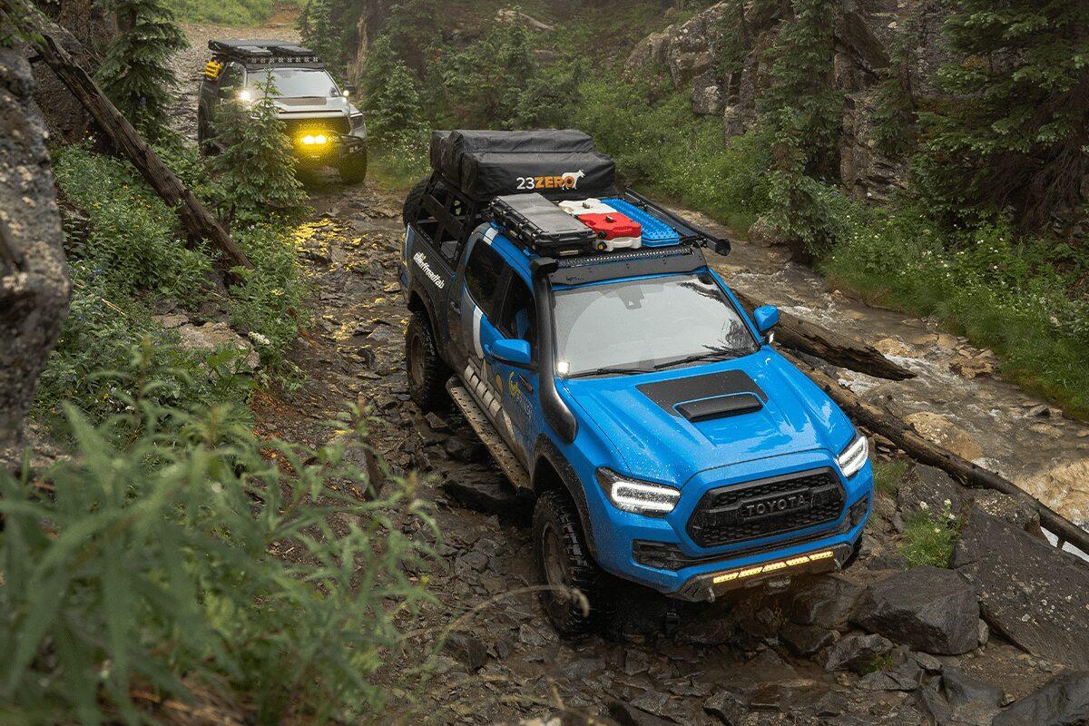 Blue Toyota Tacoma with Prinsu roof rack and camping gear off-roading on rocky forest trail