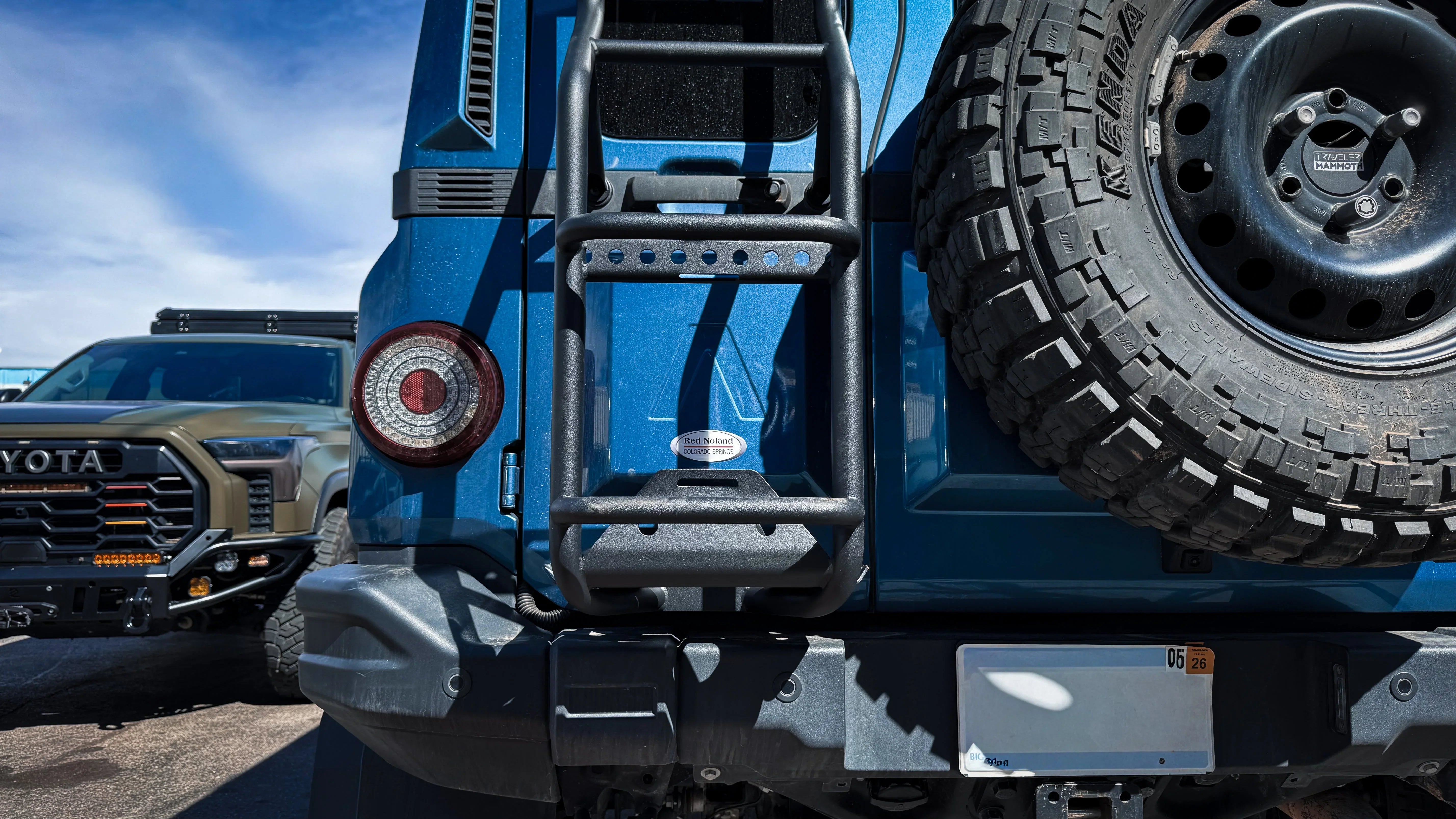 Rear ladder with anti-slip texture on blue overland vehicle, spare tire and Toyota truck visible