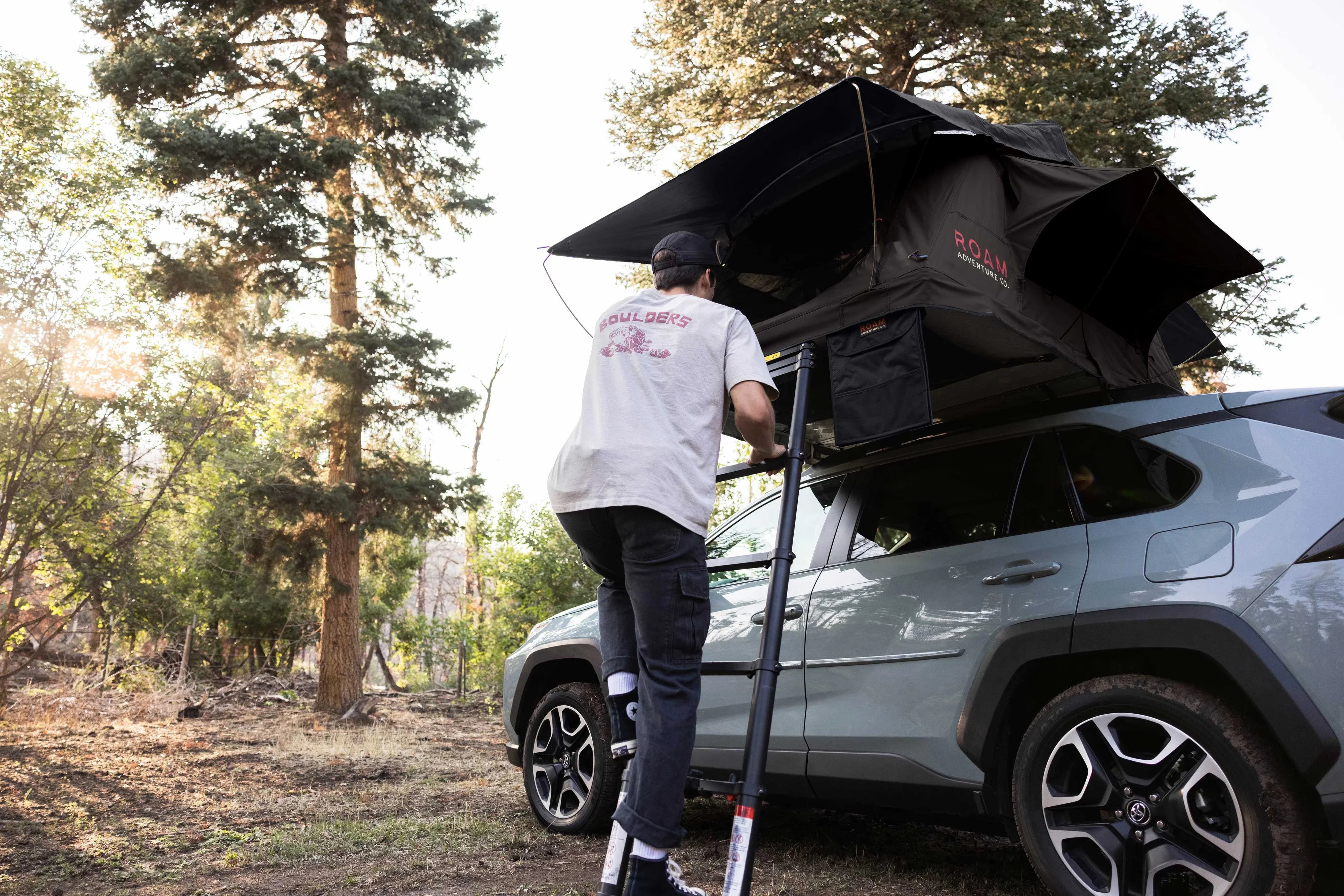 Man climbing ladder to rooftop tent on SUV in forest, overlanding camping gear scene