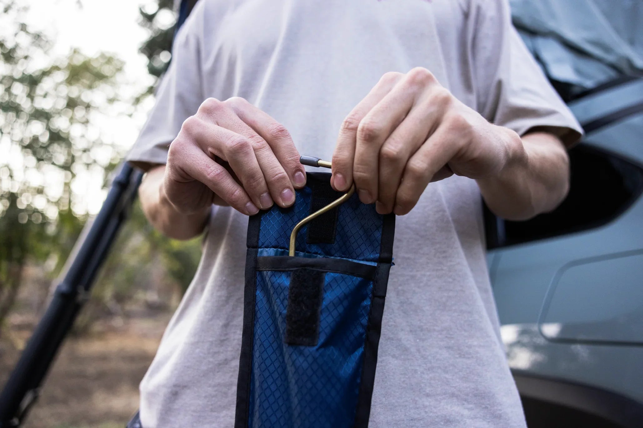 Person inserting a metal camping tool into a blue pouch near an off-road vehicle outdoors