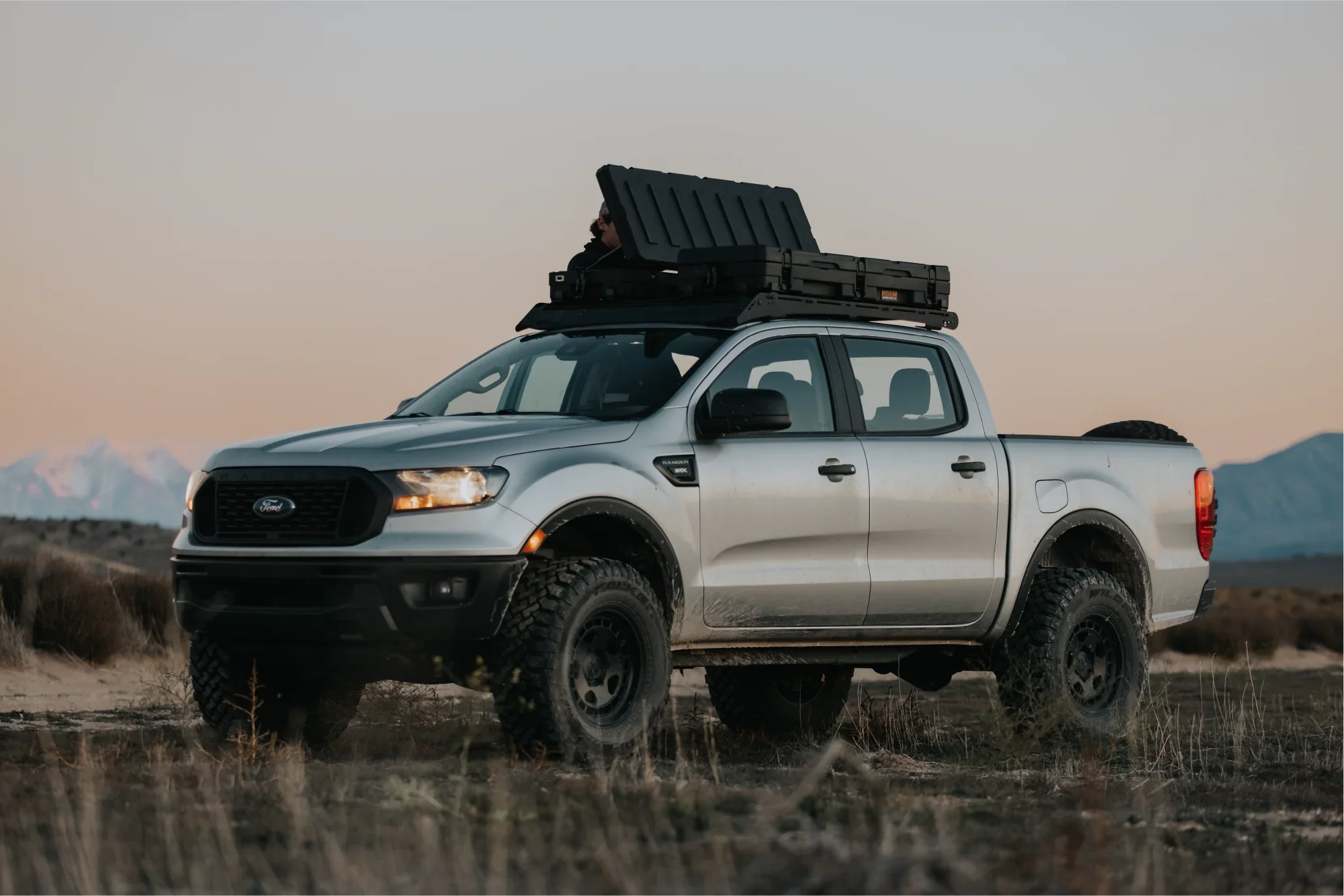Off-road pickup truck with rugged roof rack storage case in a wilderness setting at sunset