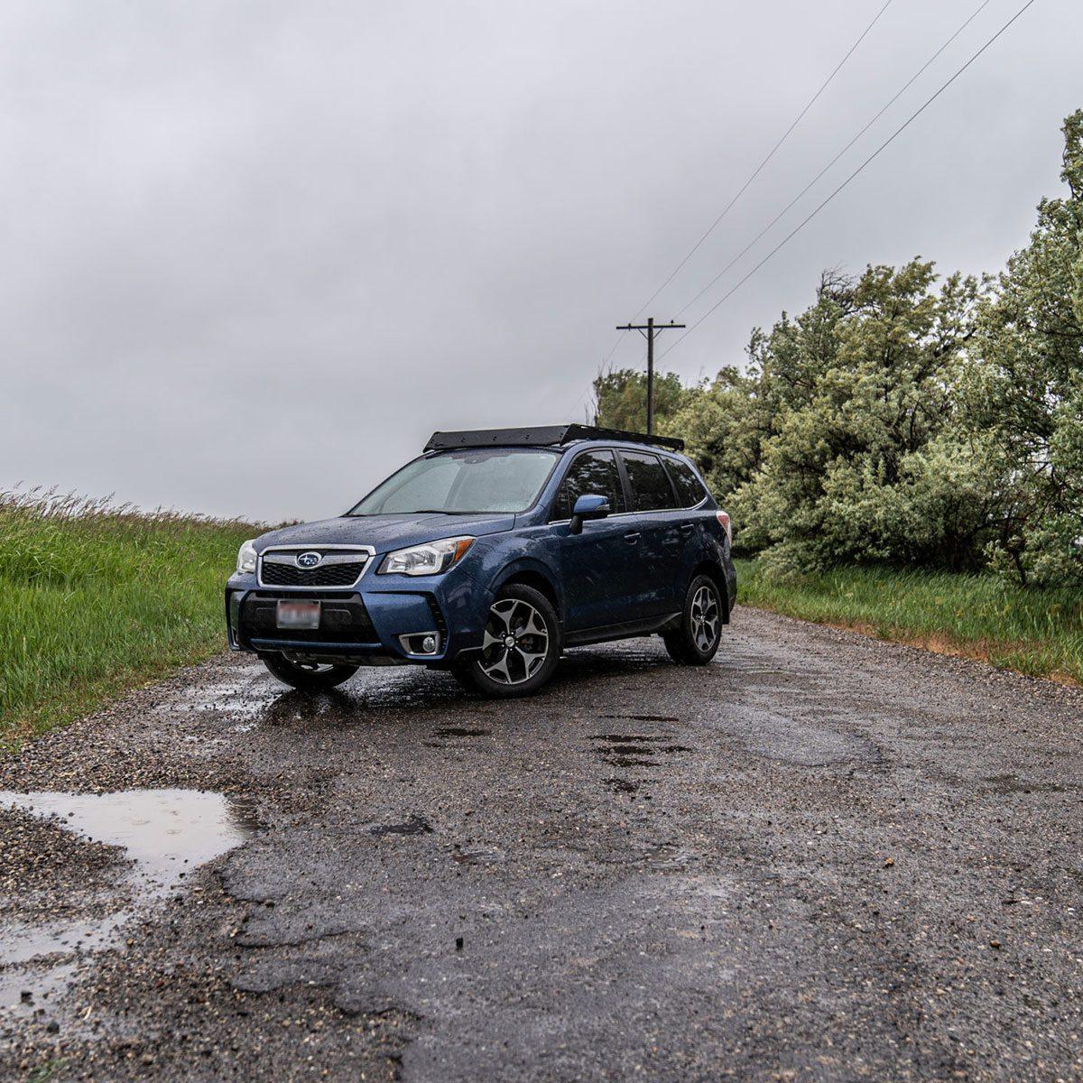 Blue Subaru Forester with roof rack parked on a wet gravel road near green trees