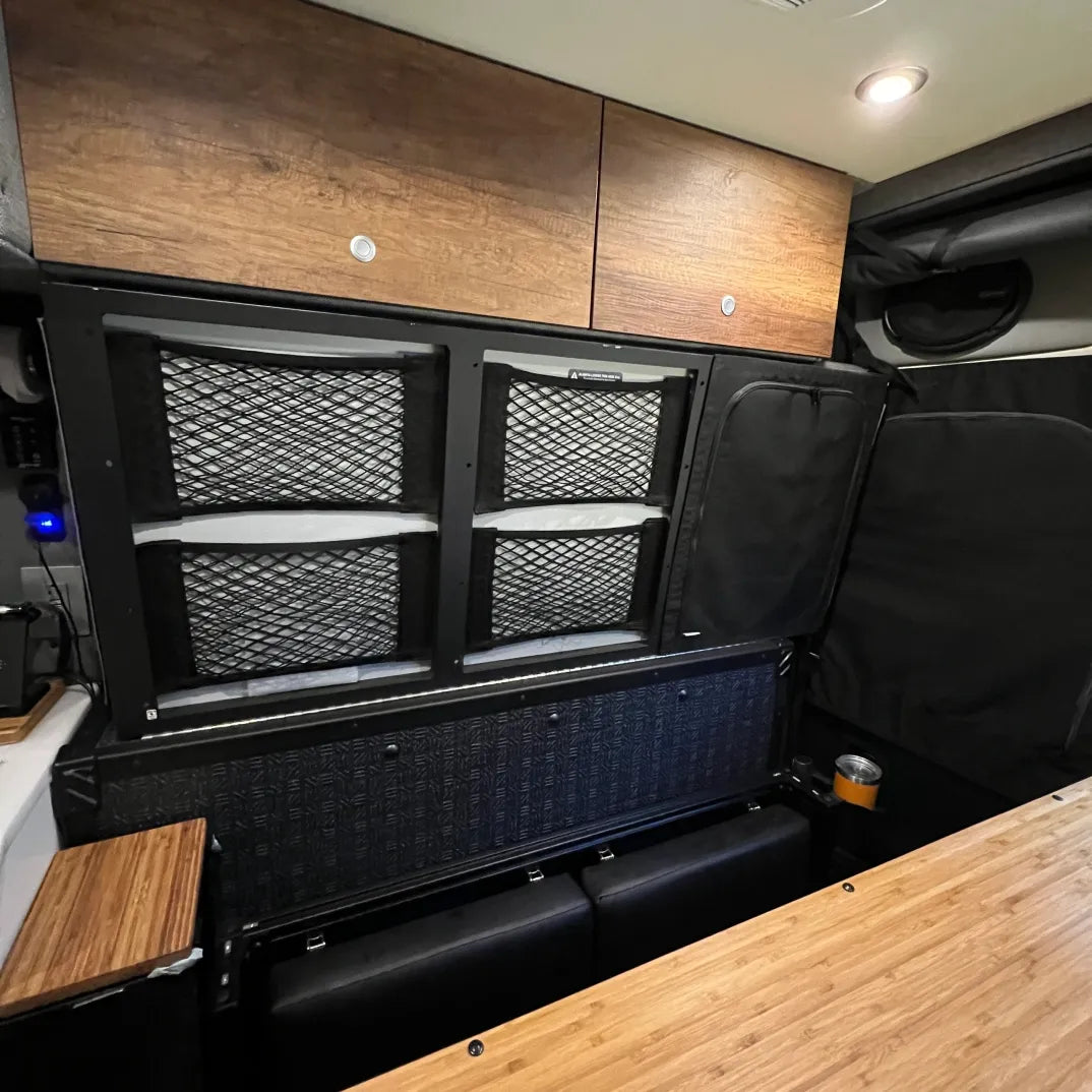 Overland vehicle interior with mesh storage panels, wood cabinets, and camping gear.