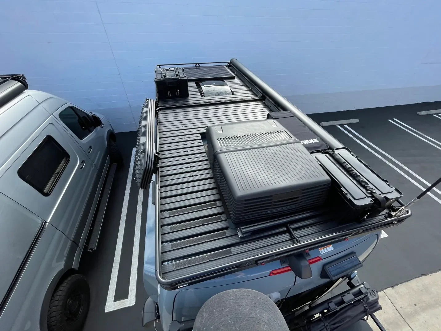 Overland vehicle roof rack with gear boxes, awning, and ladder in a parking lot