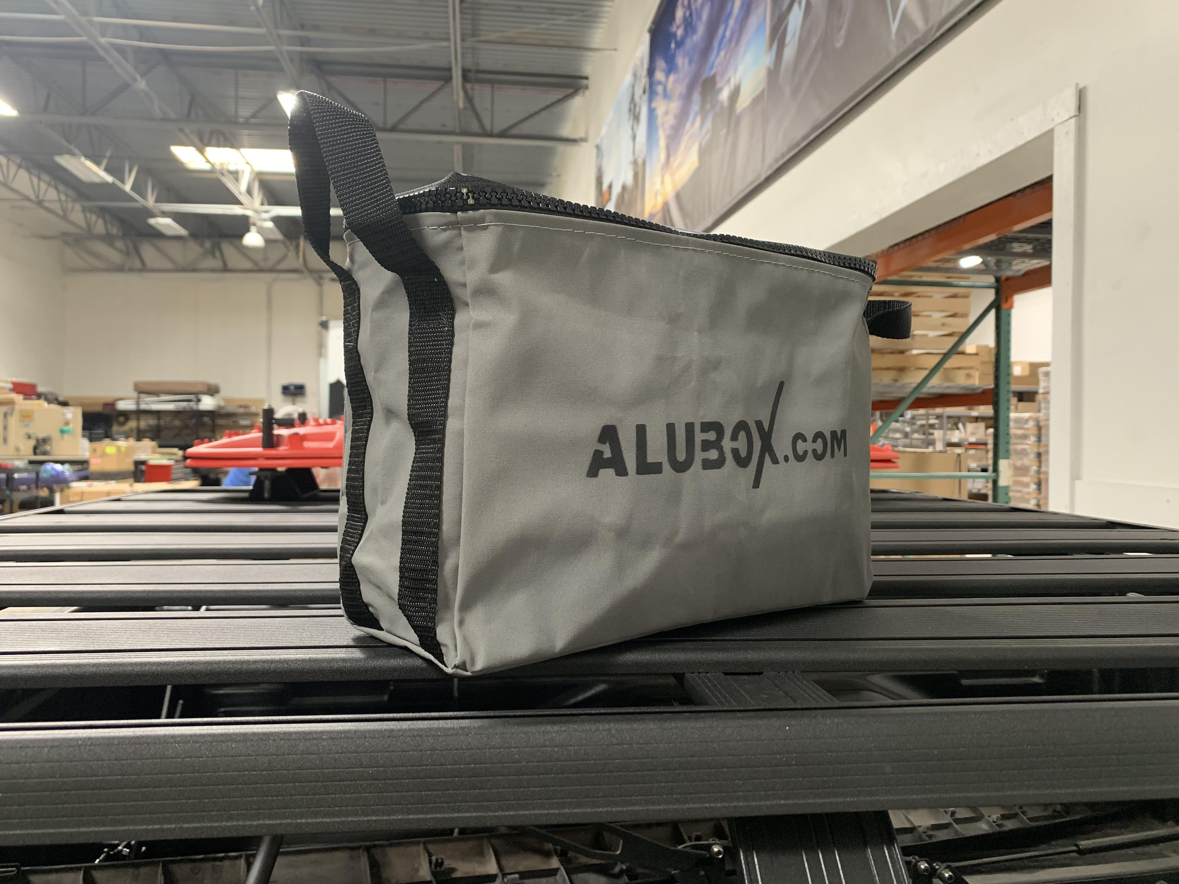 Gray ALUBOX.com gear bag on a vehicle roof rack in a warehouse setting, overlanding accessory